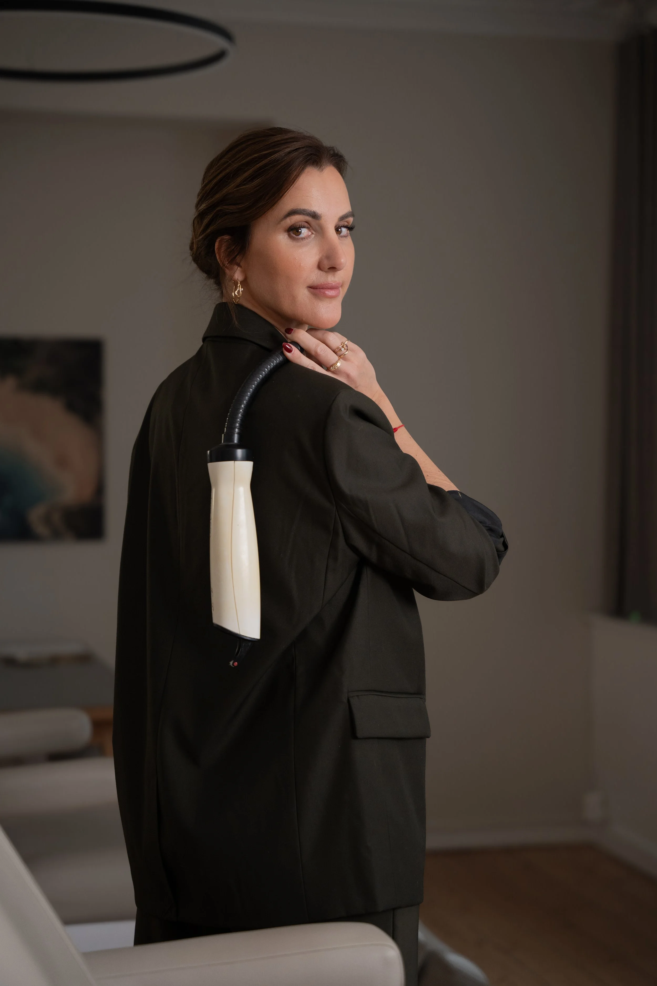 Kvindelig person med brunt hår, iført sort jakke, står i et rum med moderne indretning, med en medicinsk enhed eller lignende over skulderen.