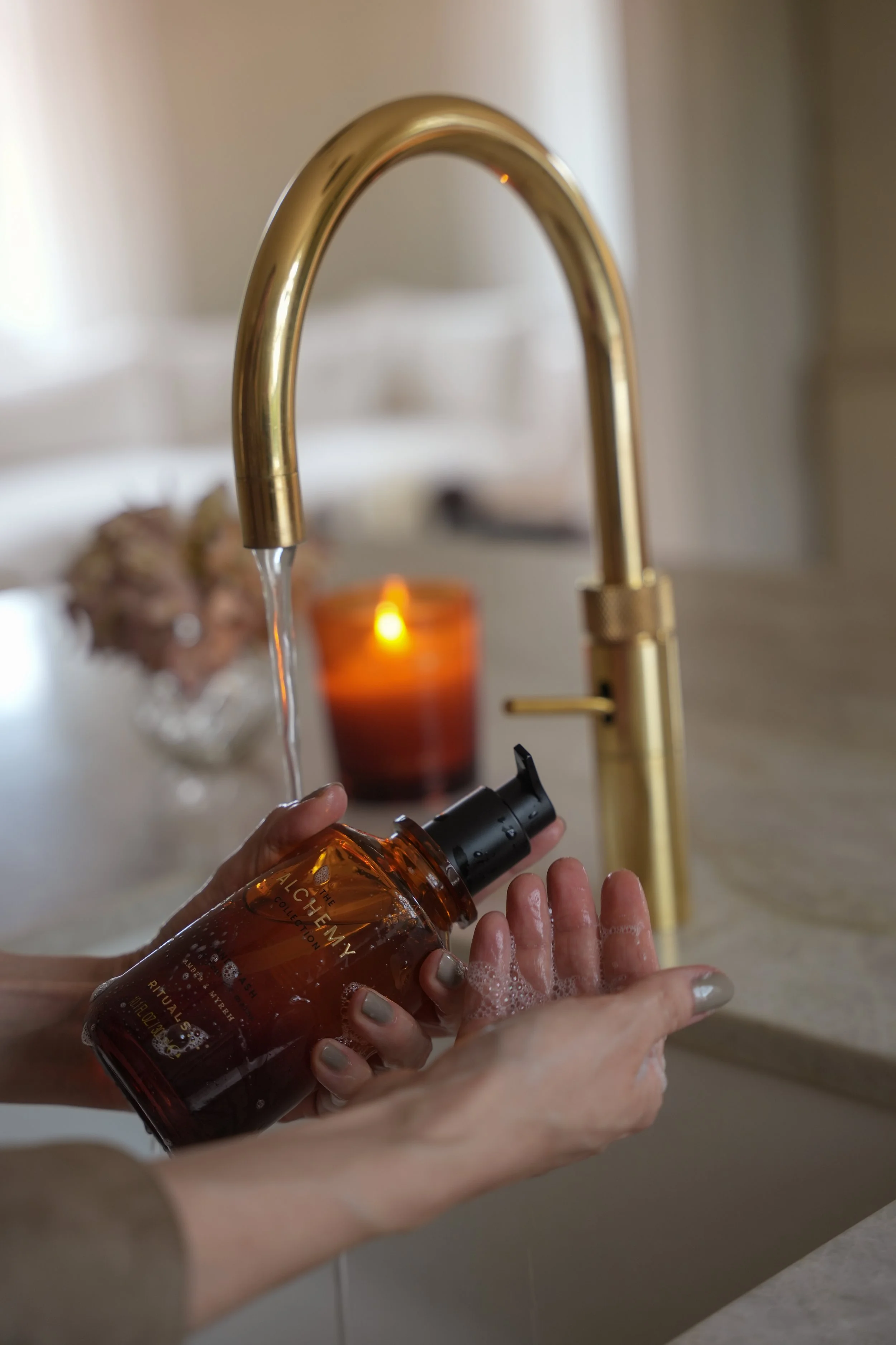 Person, der vasker hænder under en gylden køkkenhane, med sæbebruser og bobler, i baggrunden en brændende lysestage og blomster.