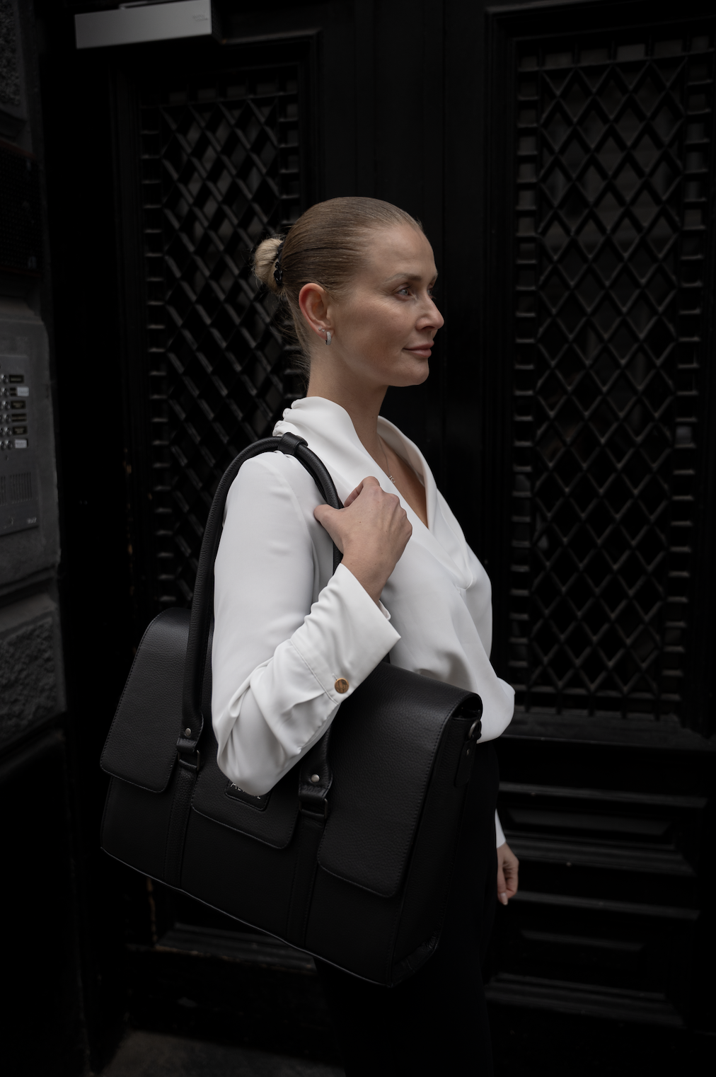 Kvindelig person iført hvid skjorte, med sort taske over skulderen, stående foran sorte porte.
