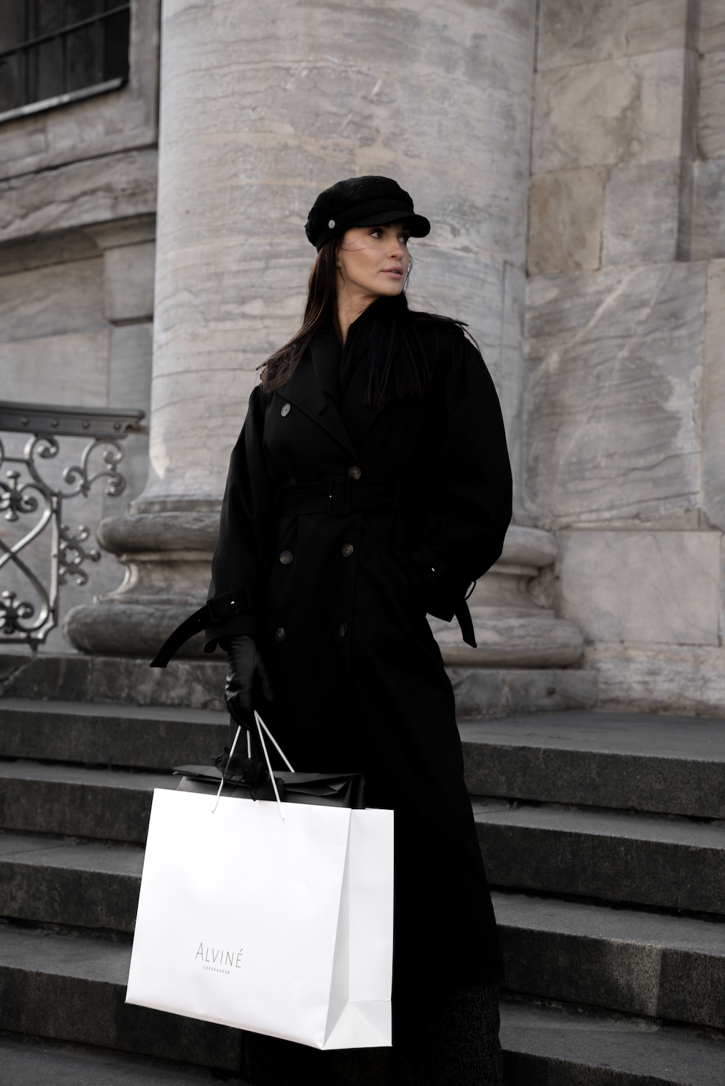 Kvinde i sort trenchcoat og sort hue står på udendørs trappe med en shoppingpose fra Alviné.