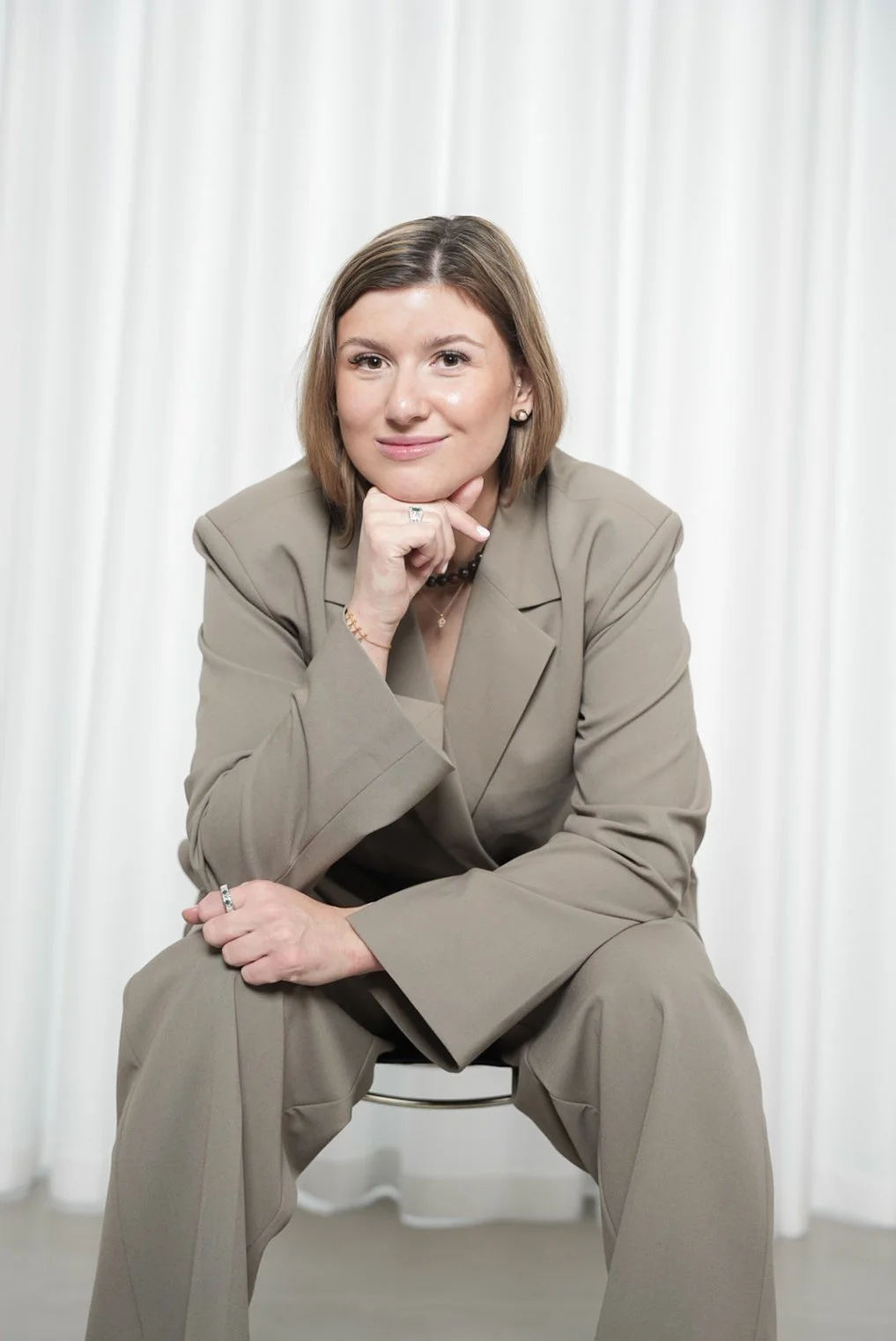 En kvinde i en beige forretningsdragt sidder på en stol foran en hvid baggrund, med håret i skulderlængde og smilende ansigt.