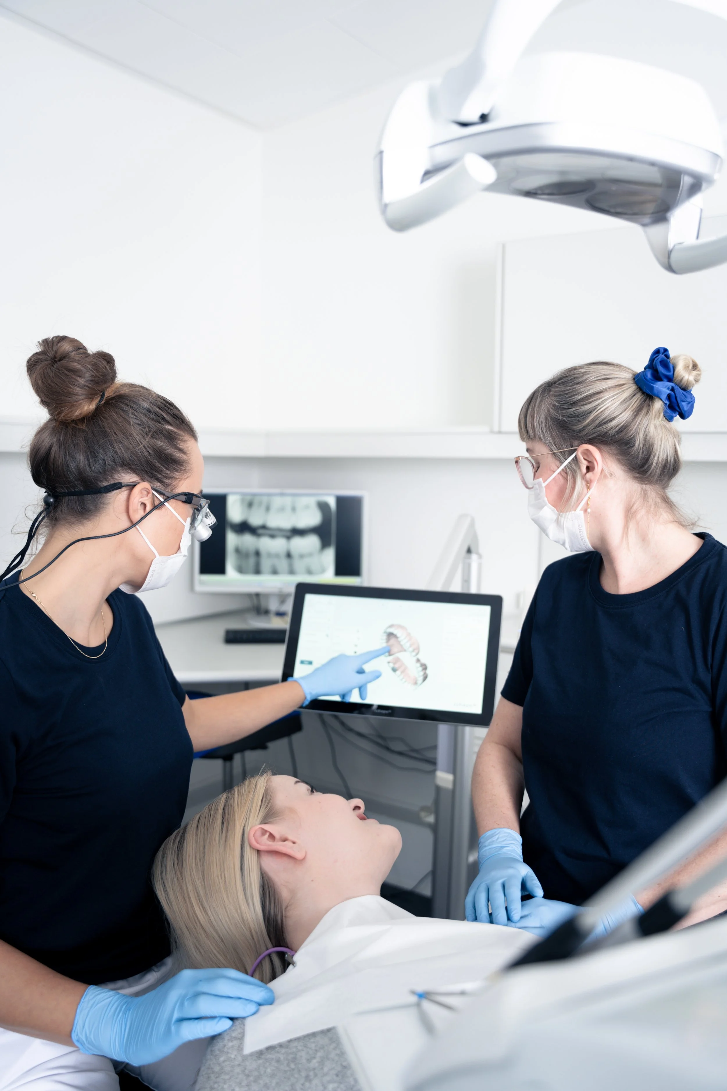 To tandlæger i maske og handsker arbejder på en patient, der ligger i en tandlægestol, mens de undersøger patientens tænder på en computerskærm.