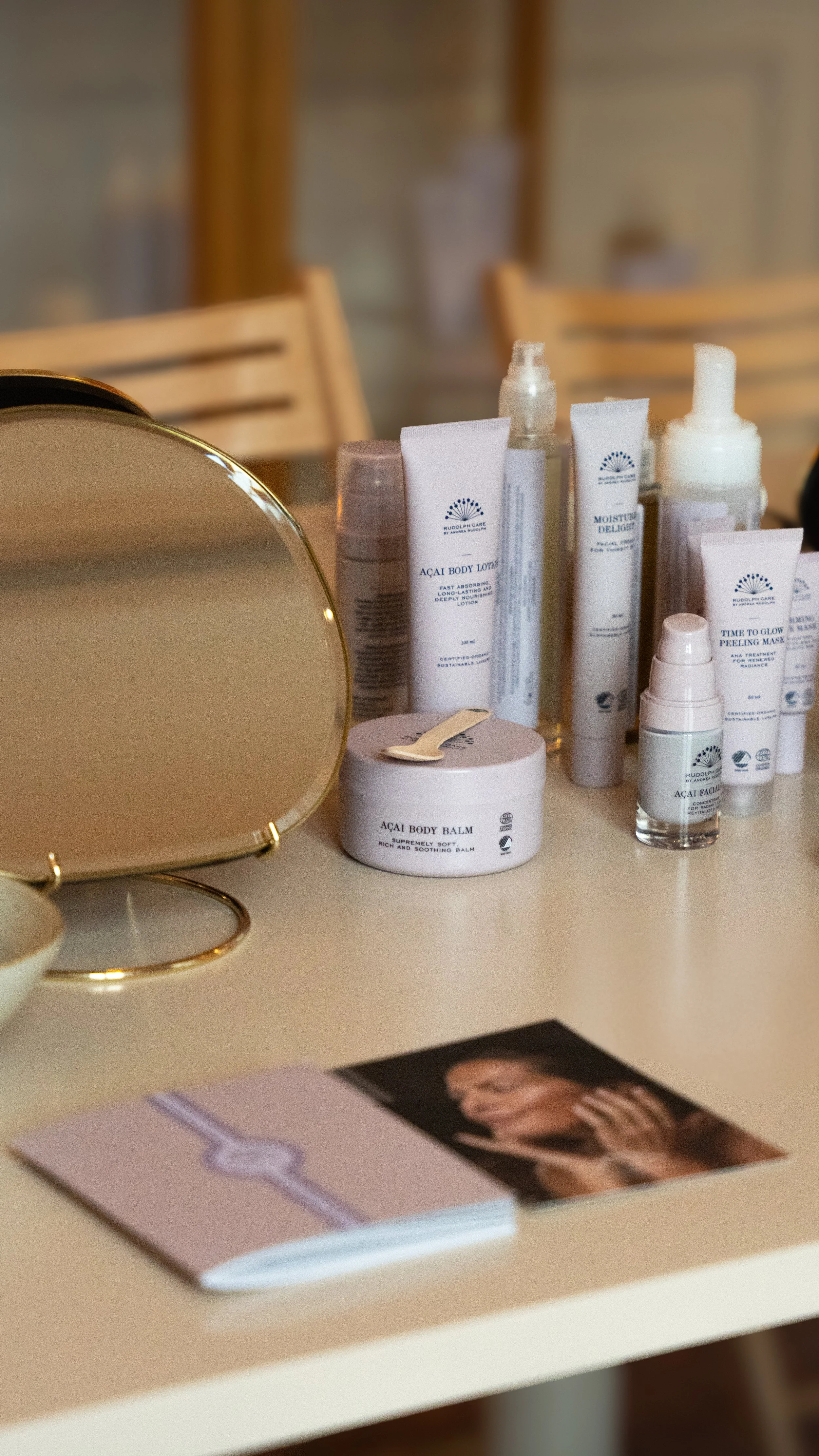 Skønhedsprodukter på et bord, herunder lotioner og cremer, med et rundt spejl og en brochure med et billede af en kvinde.