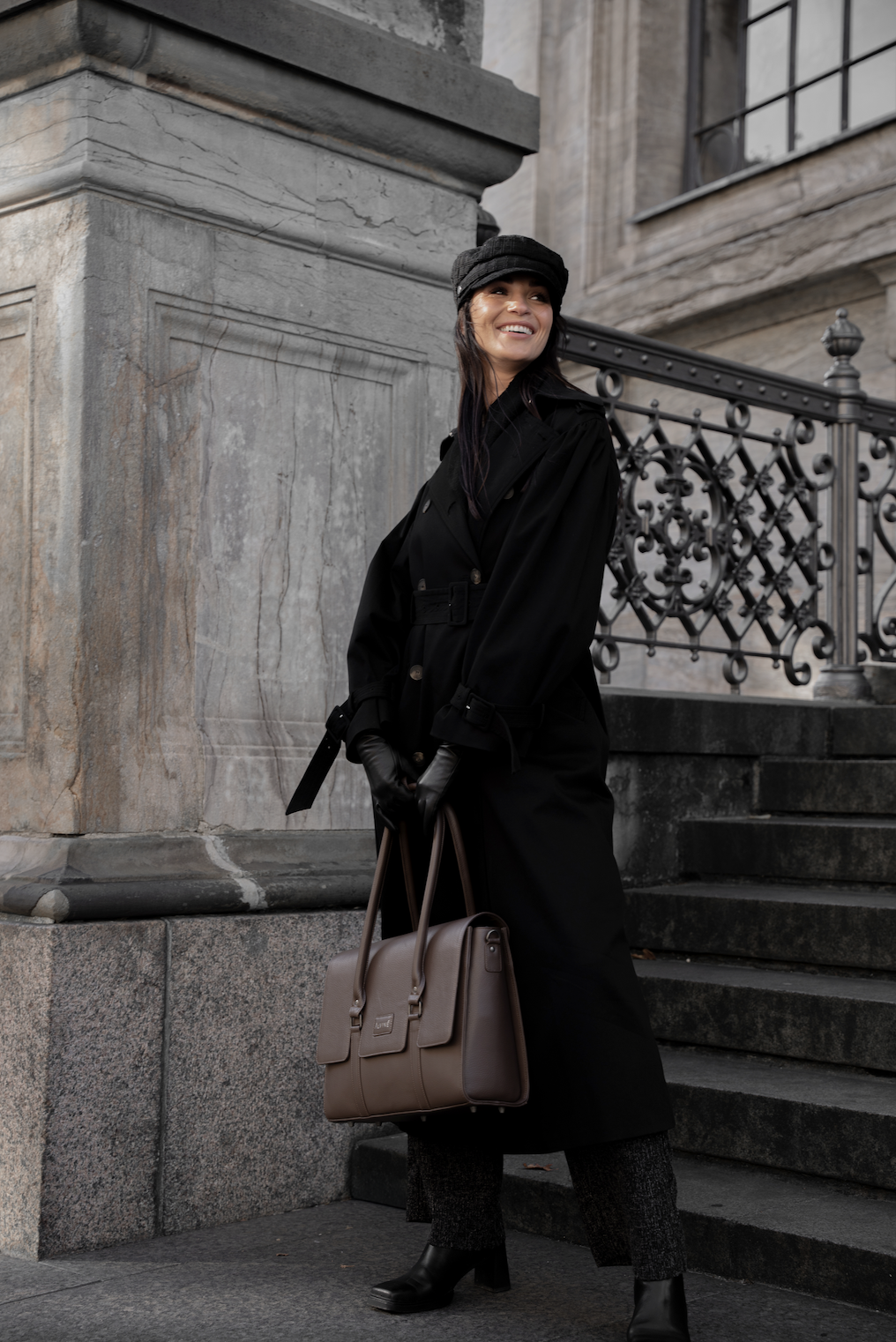 En kvinde i sort jakke, hat og handsker står udenfor ved en trappe med smedejernsgitter og en gammel bygning, hun smiler og bærer en beige taske.