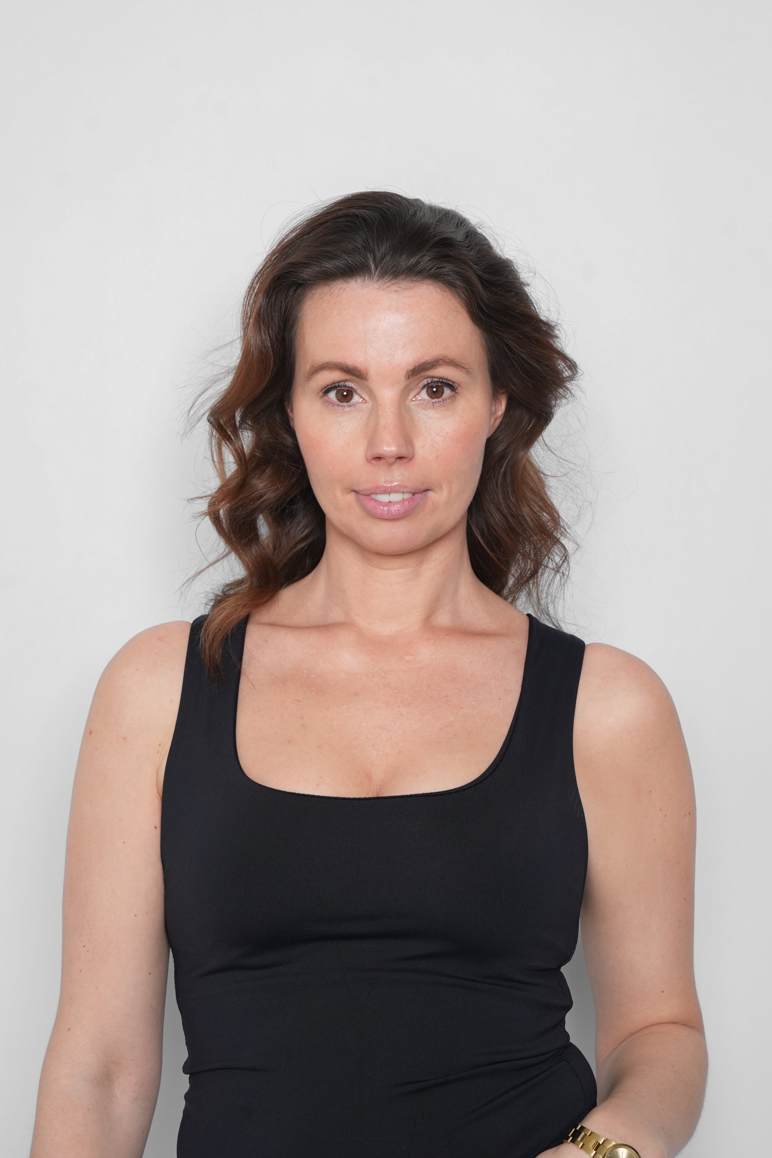 Portræt af en kvinde med mellemlangt, krøllet brunt hår, iført sort top og guldarmbånd, stående mod en neutral hvid baggrund.