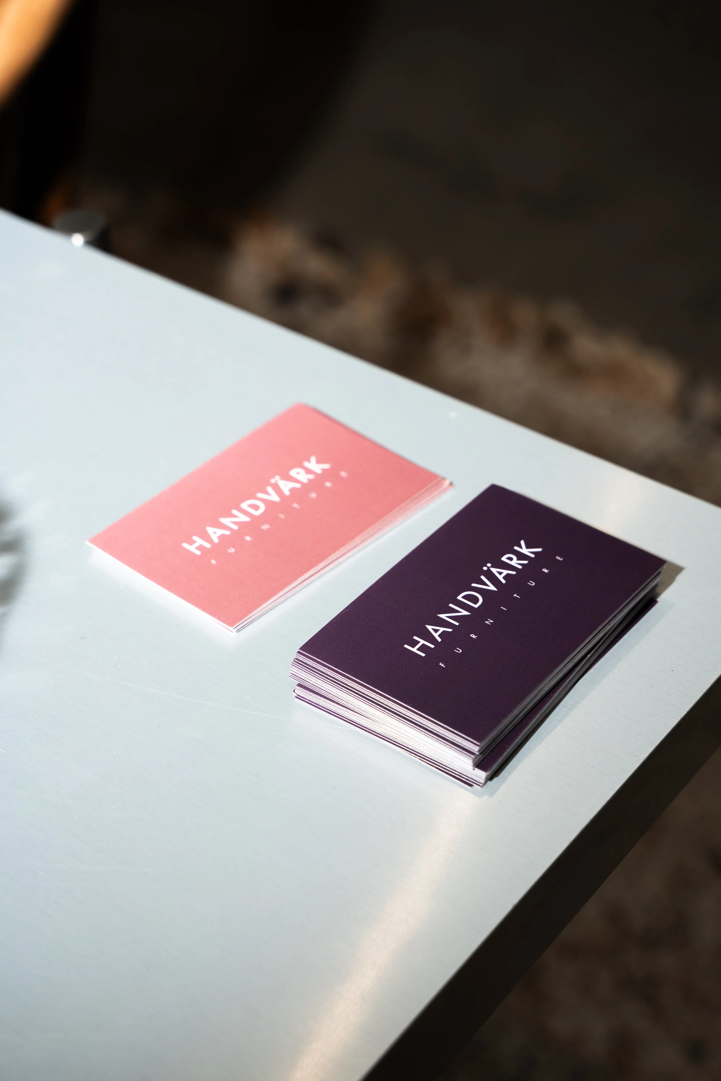 To stykker visitkort, ét pink og ét mørklød, med teksten "HANDVÆRK FURNITURE" på begge, ligger på en hvid overflade.