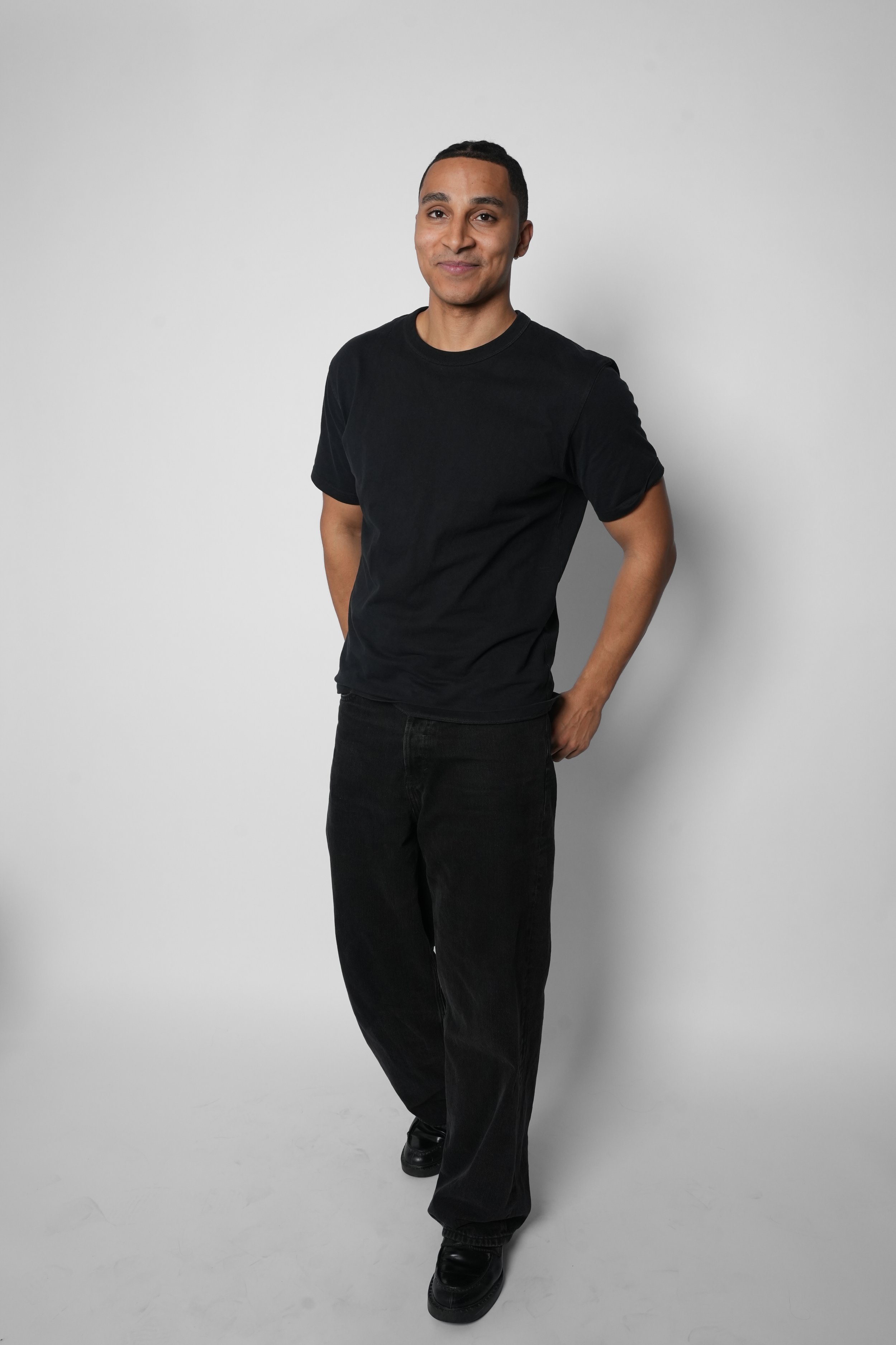 Mand står foran en hvid baggrund, iført sort t-shirt og sorte bukser, smilende med hænder i lommer.