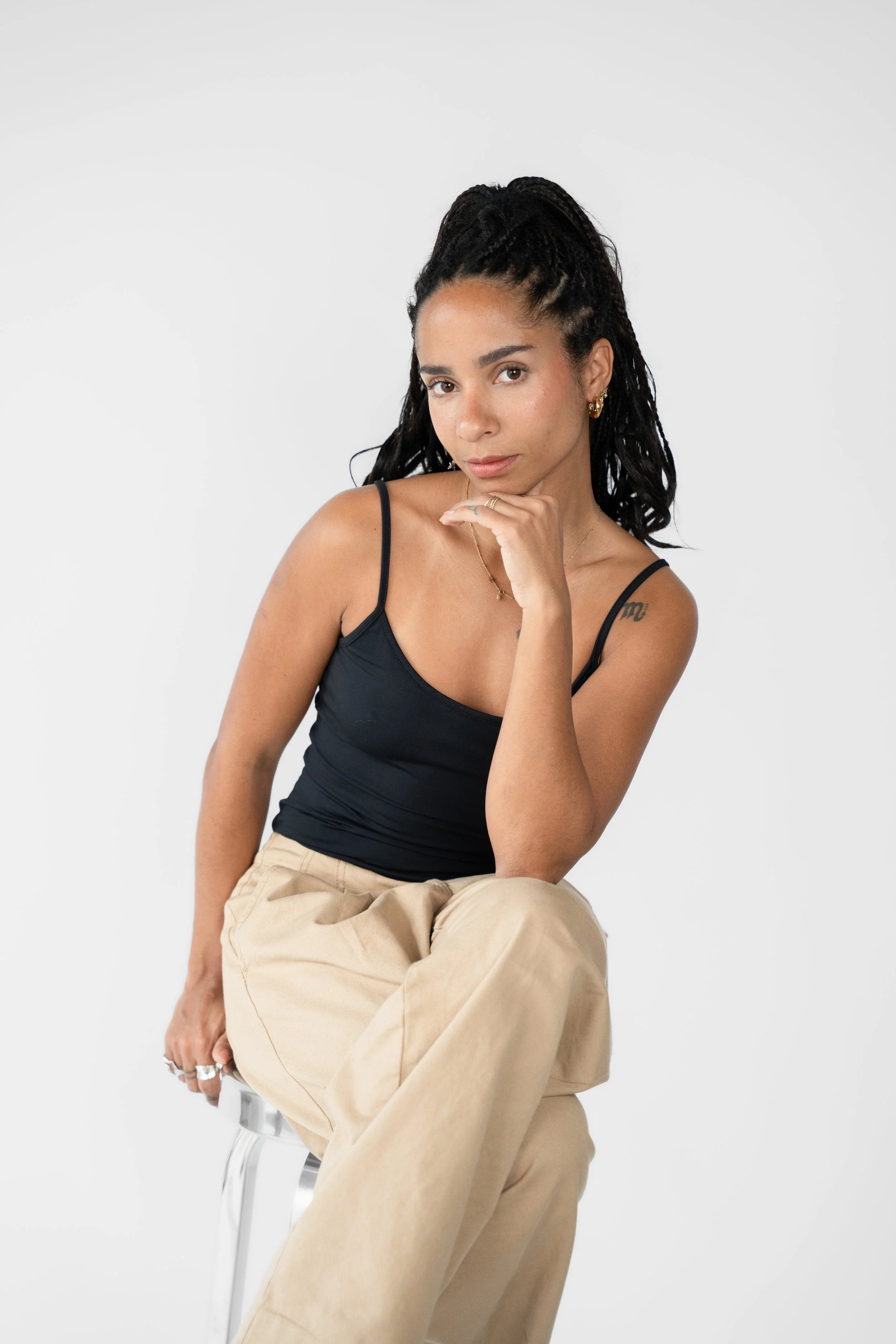 Kvinde med sort, flettet hår, iført sort top og beige bukser, sidder på en gennemsigtig stol mod en hvid baggrund.