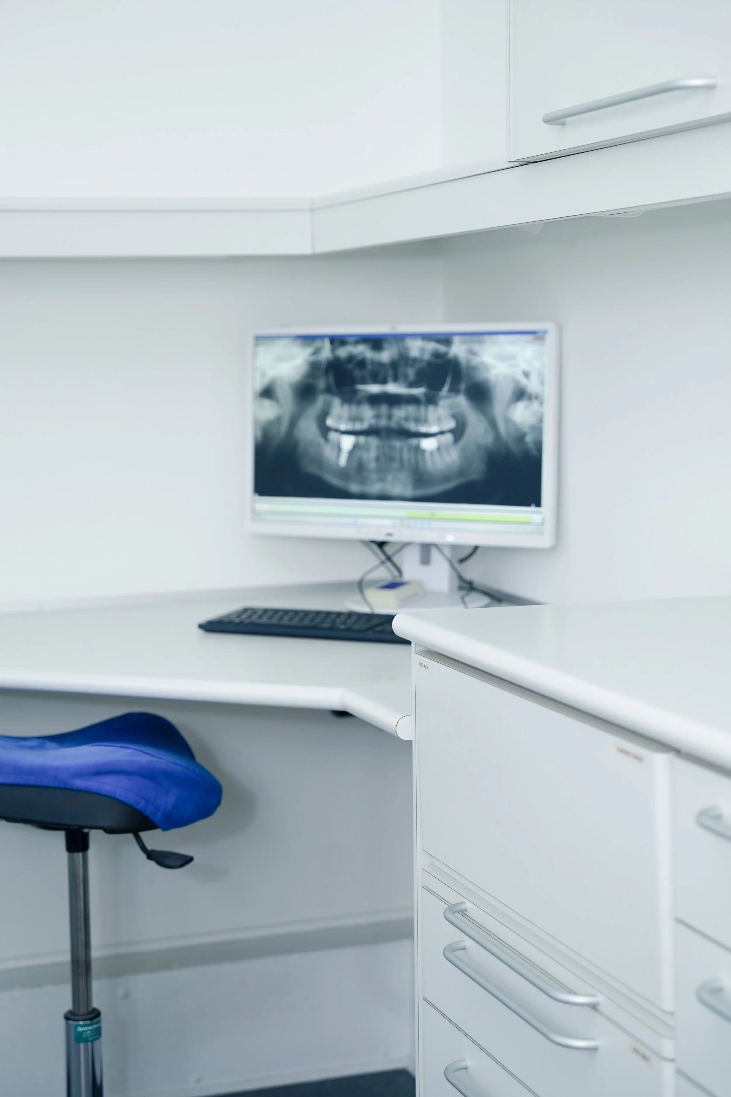 Et foto af et læge- eller klinikkontor med en computerskærm, der viser en tand- eller kæbex-ray. Der er en kontorstol og nogle skuffer. Væggene er hvide.