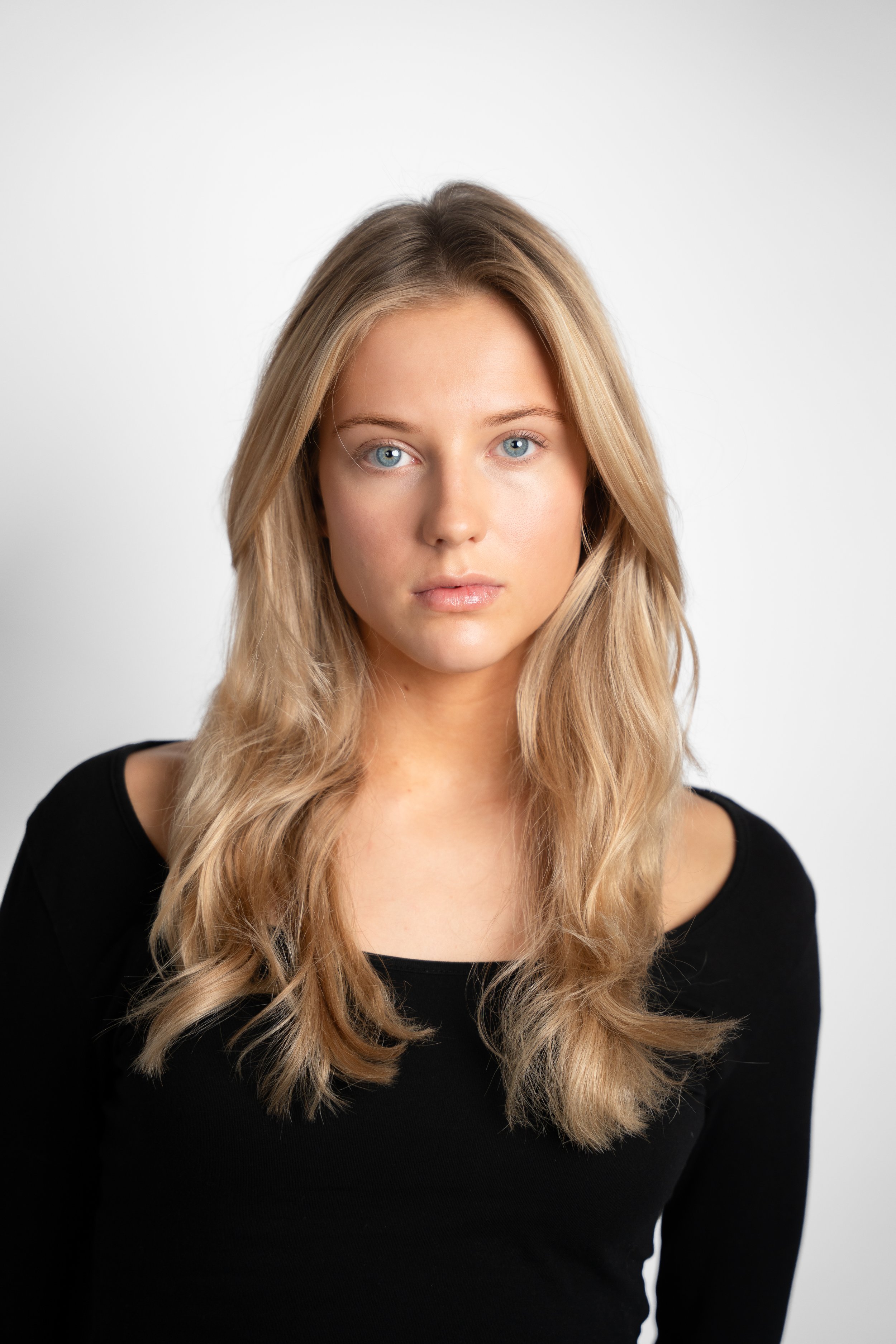 Portræt af en ung kvinde med blond hår og blå øjne iført sort tøj, stående mod en hvid baggrund.