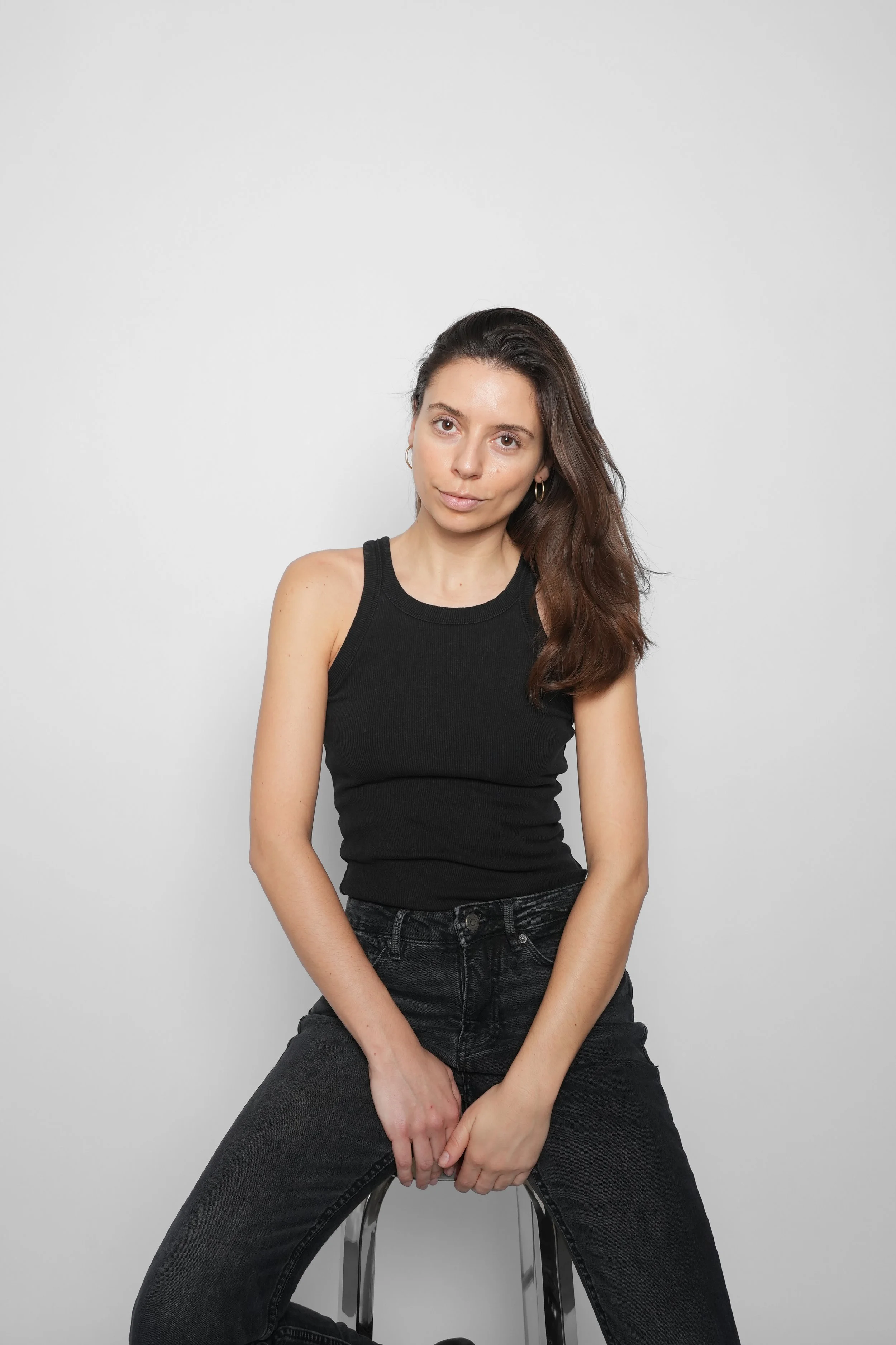 En kvinde sidder på en stol foran en hvid baggrund, iført sort top og jeans.