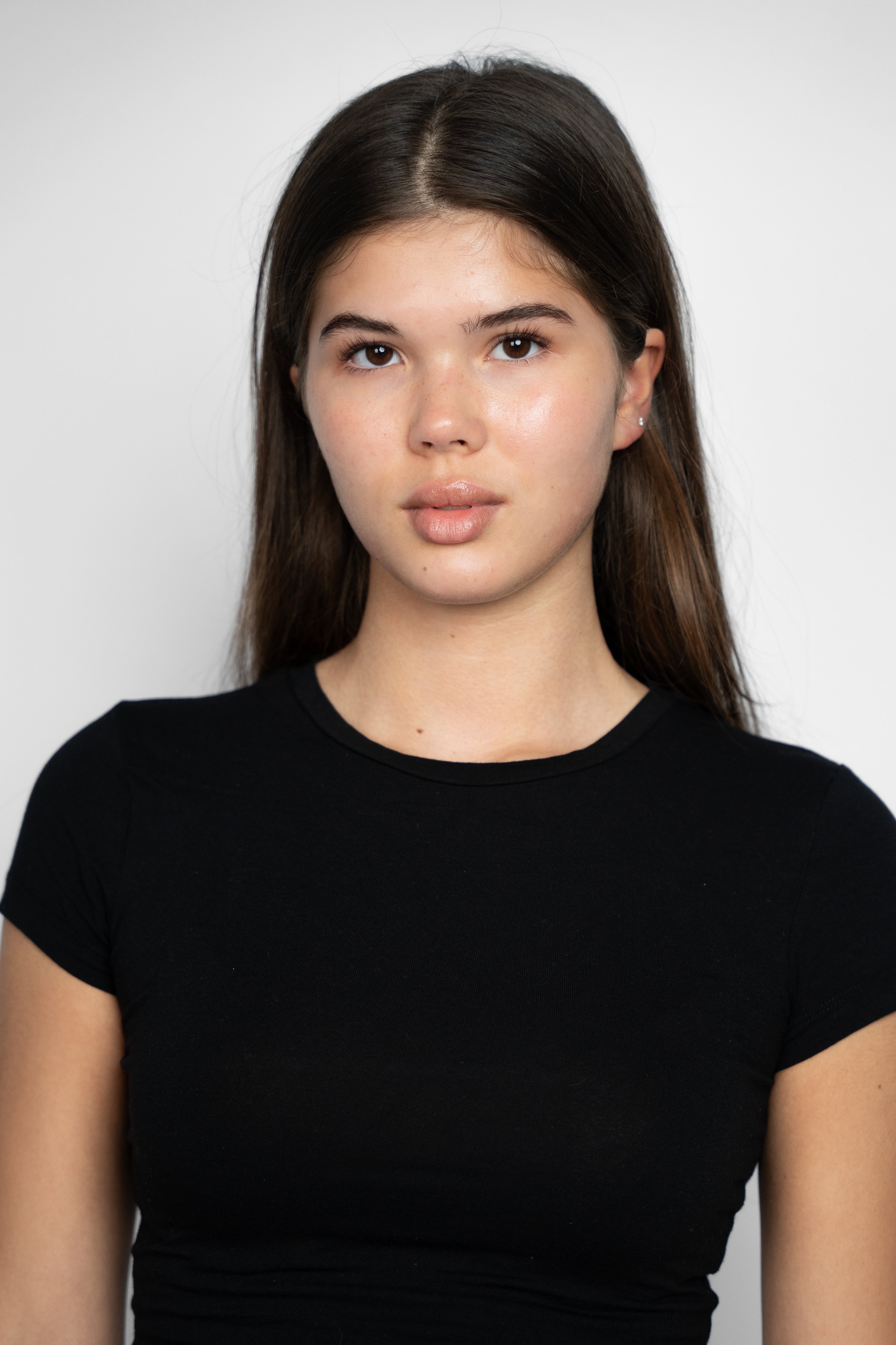 Portræt af en ung kvinde med langt brunt hår og let makeup, iført en sort t-shirt, stående foran en hvid baggrund.