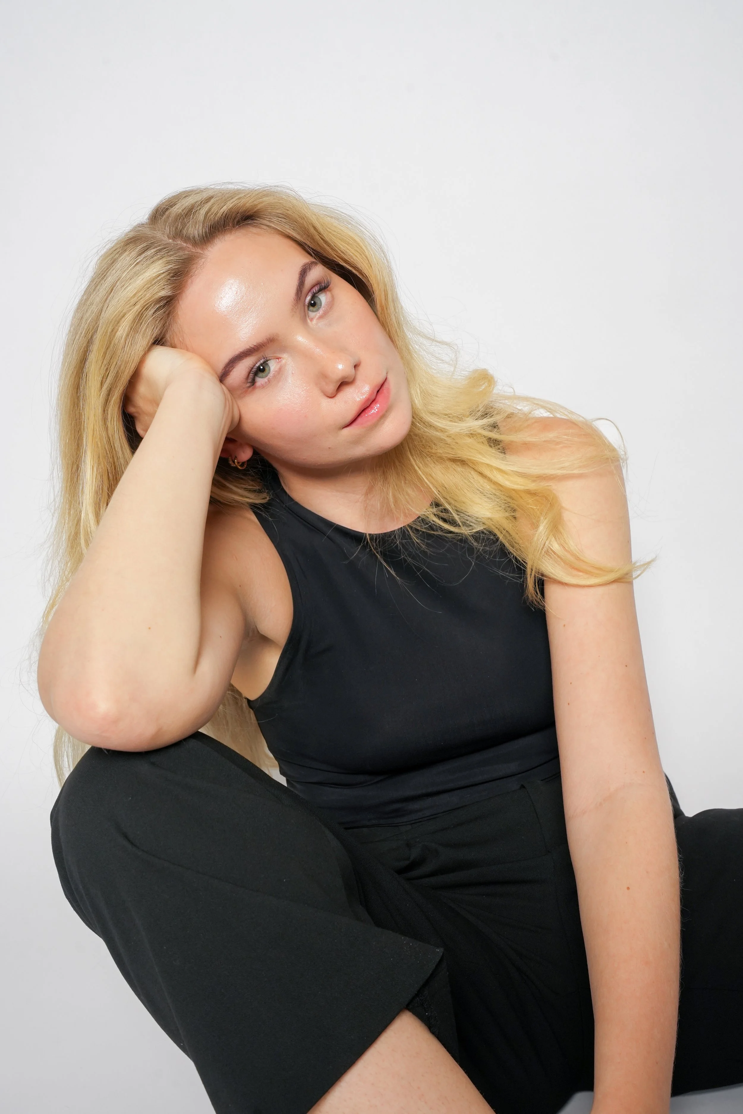 Portræt af en ung kvinde med blondt hår, iført sort top og bukser, siddende med hovedet støttet af hånden mod kinden, mod en hvid baggrund.