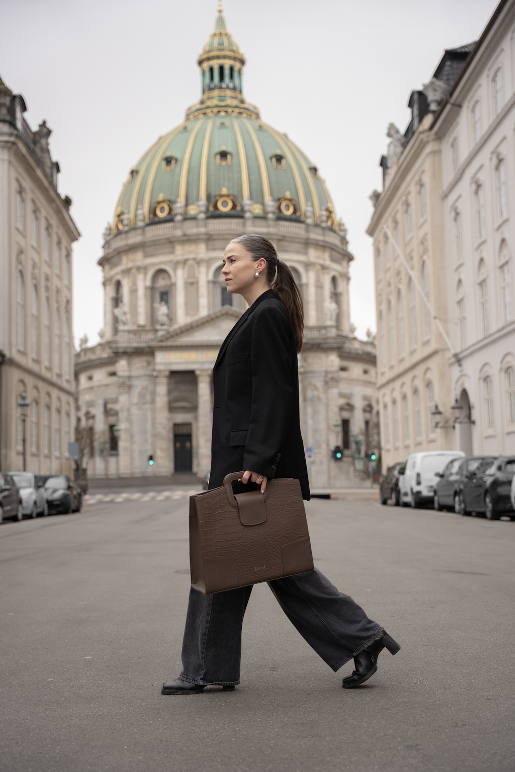 En person i sorte tøj, der går over en bygade med en stor kupolbygning i baggrunden, og bærer en brun taske.