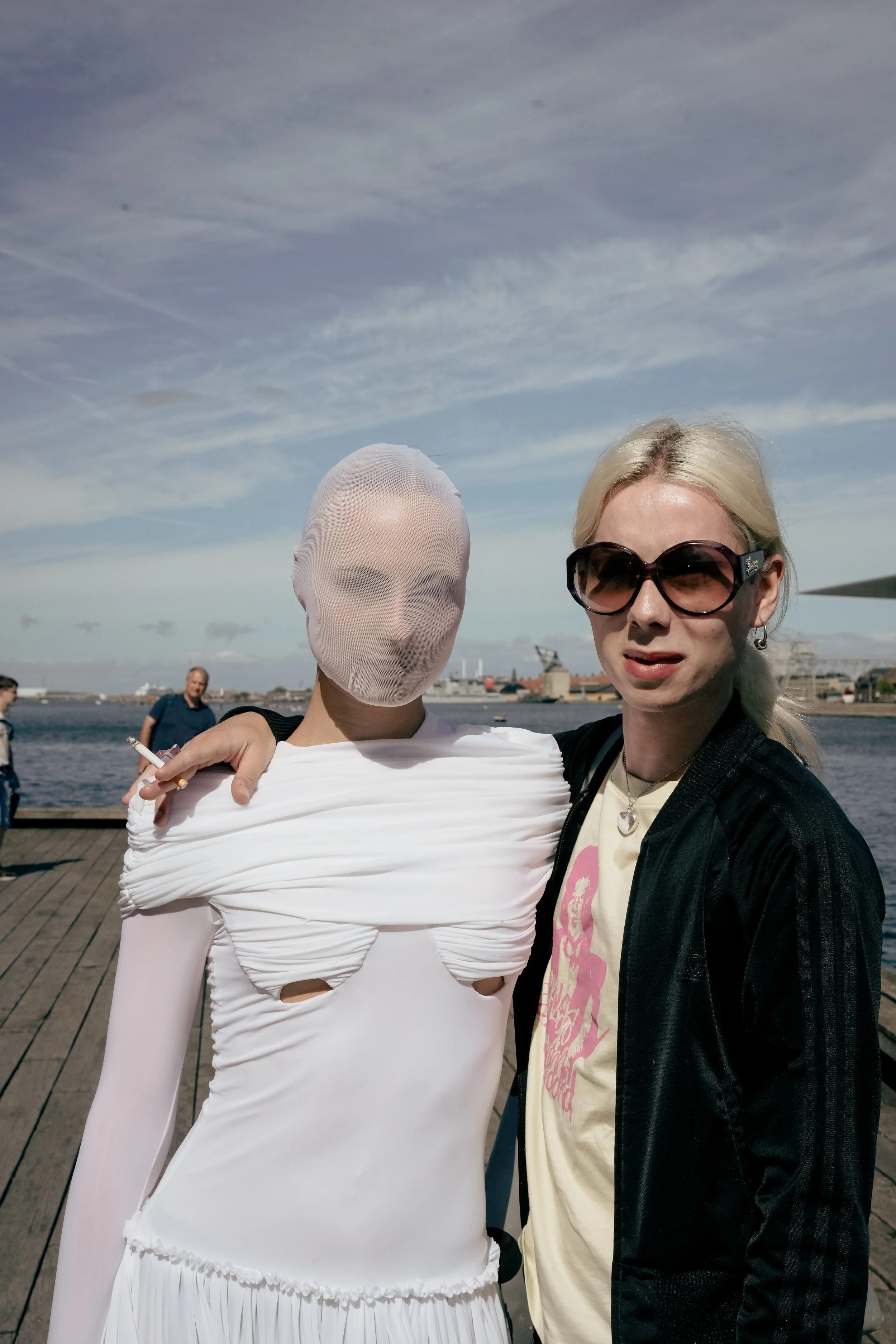 To kvinder står tæt sammen udendørs ved vandet, den ene med mundbind og den anden med store solbriller og blondt hår, med by og skibe i baggrunden.