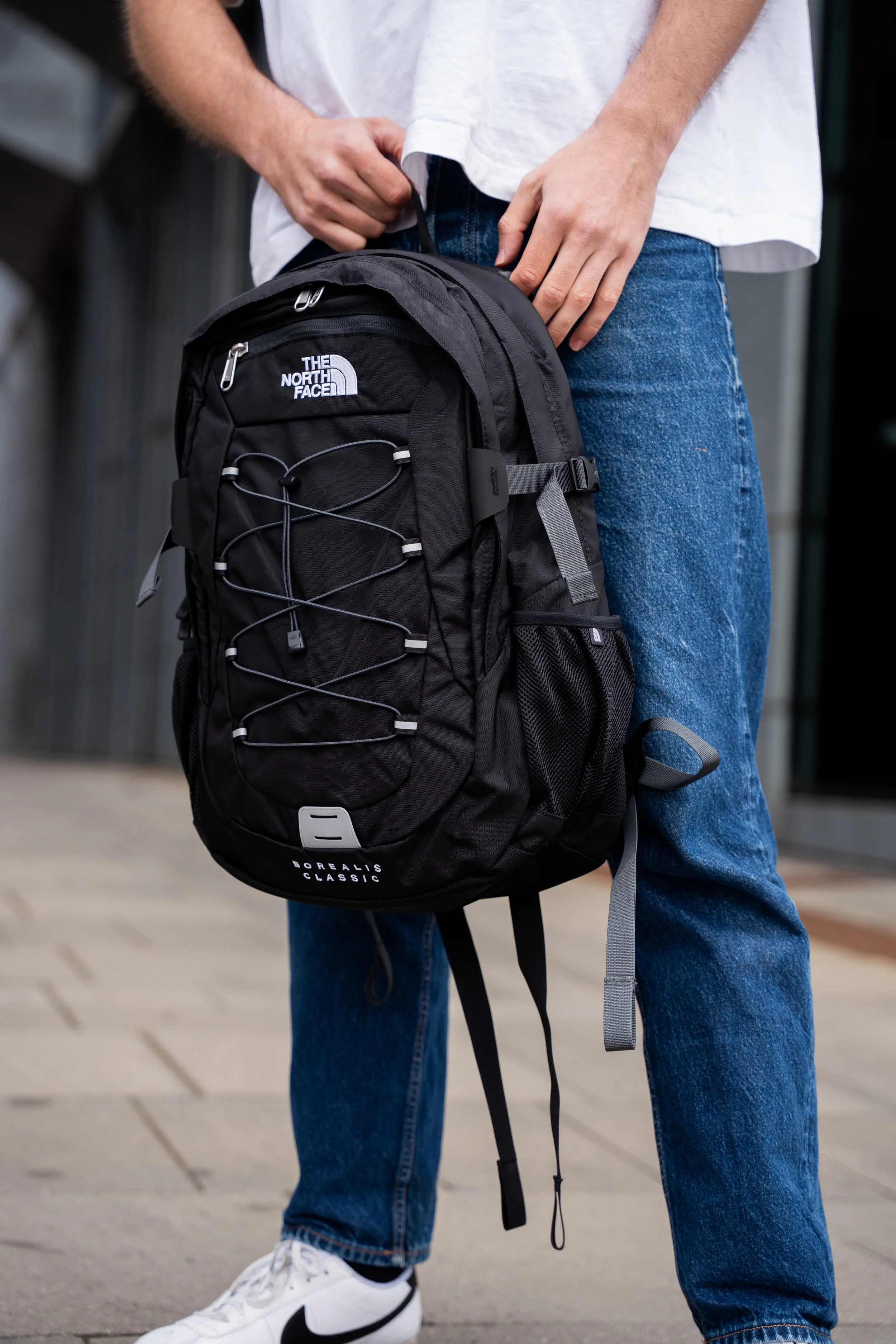 Person, der står med en sort The North Face rygsæk, iført blå jeans og hvide sneakers.