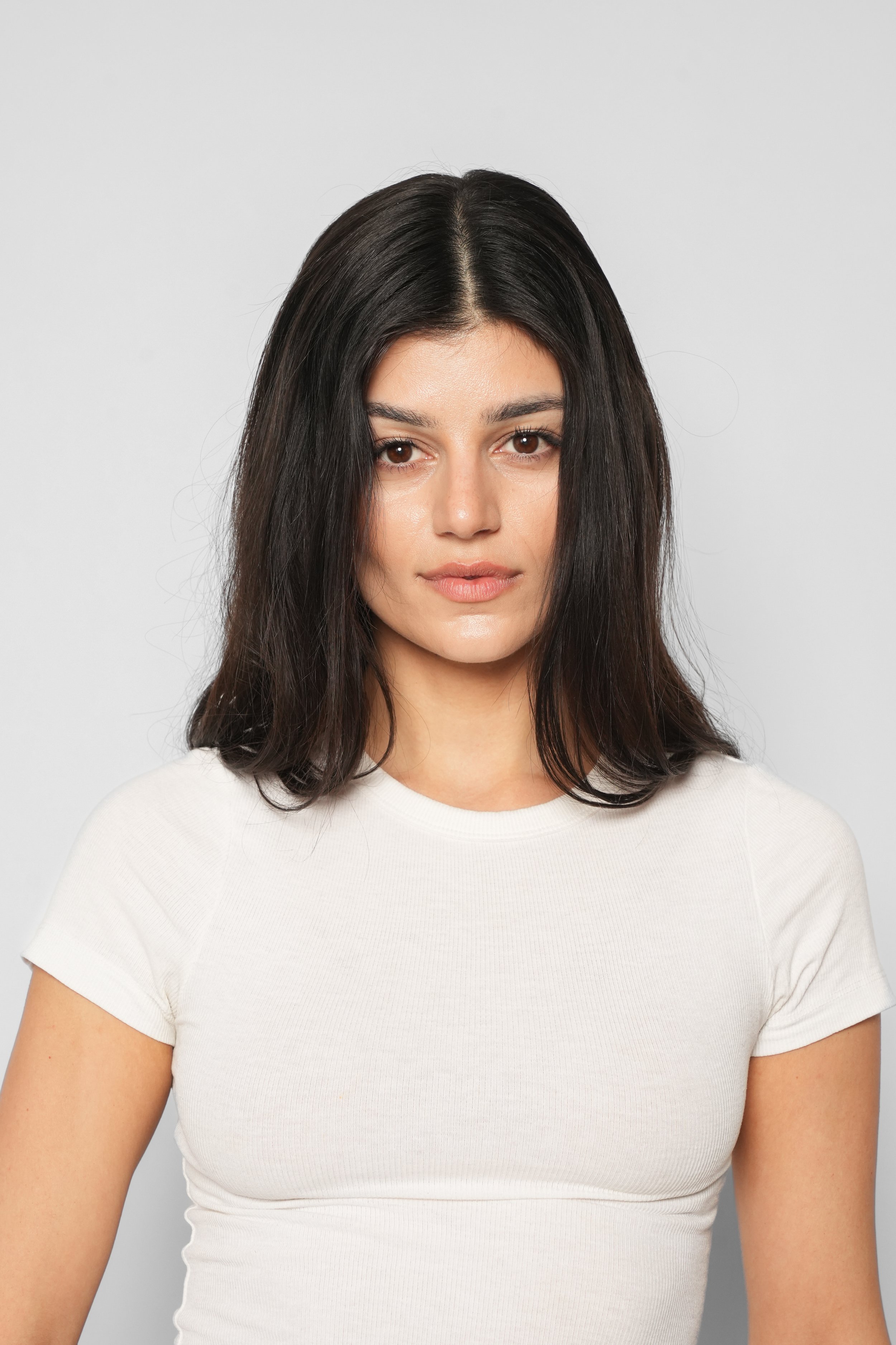 Portræt af en ung kvinde med skulderlangt sort, blankt hår, iført hvid t-shirt, mod en lys grå baggrund.