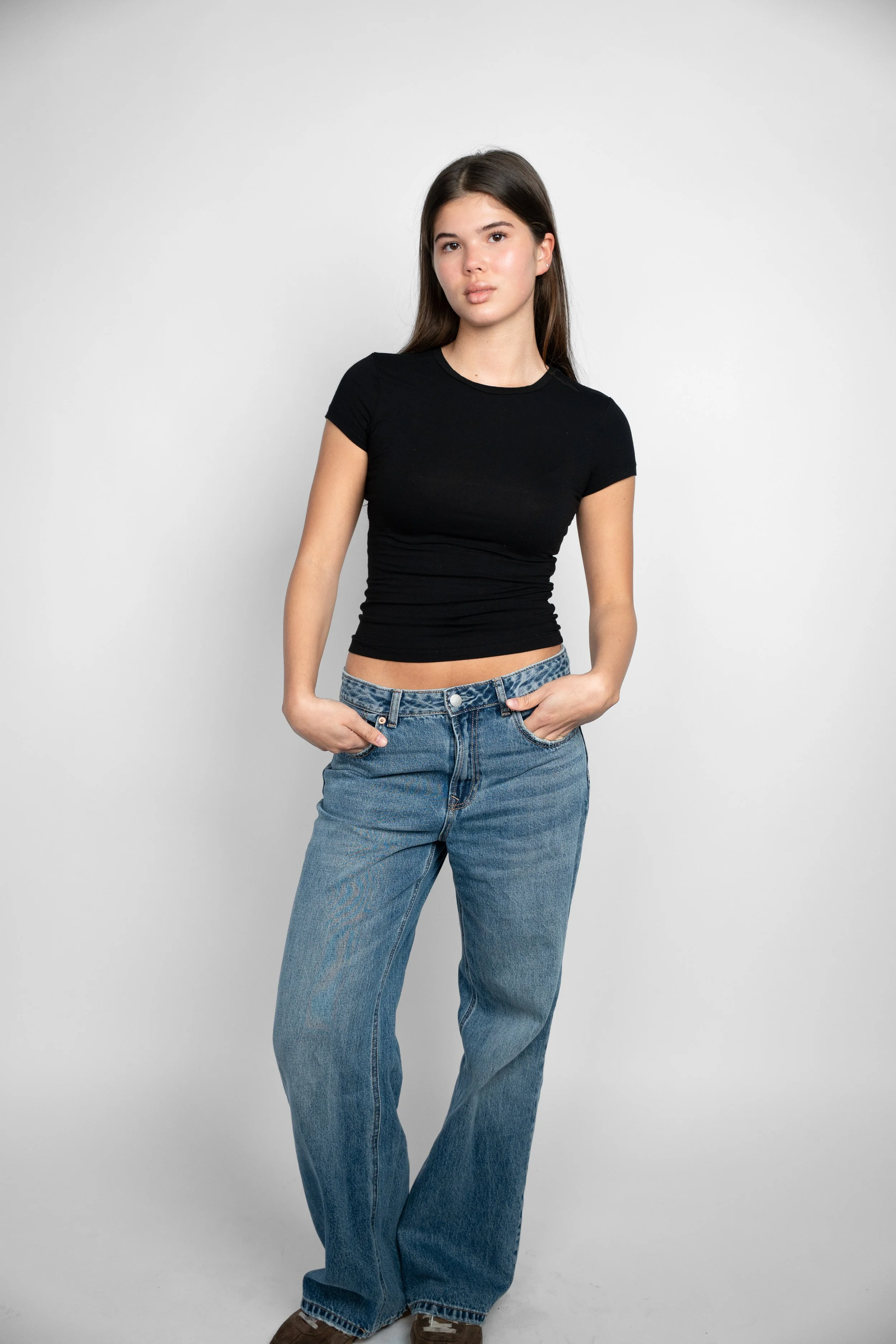 Ung kvinde med langt brunt hår, iført sort t-shirt og blåjeans, står foran en hvid baggrund.