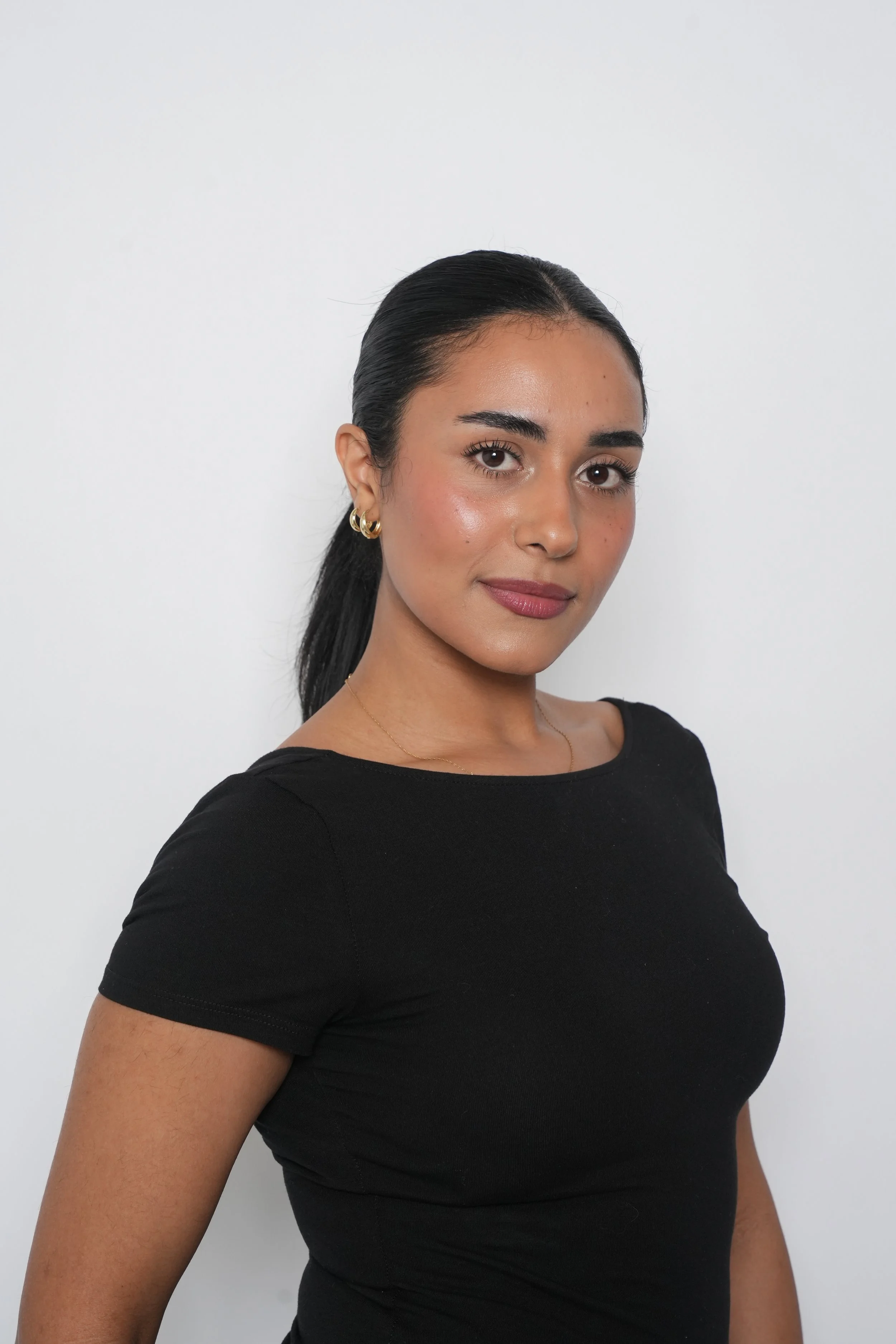 Portræt af en ung kvinde med sort hår, iført sort t-shirt, satellithalskæde og guldhalskæde mod en hvid baggrund.