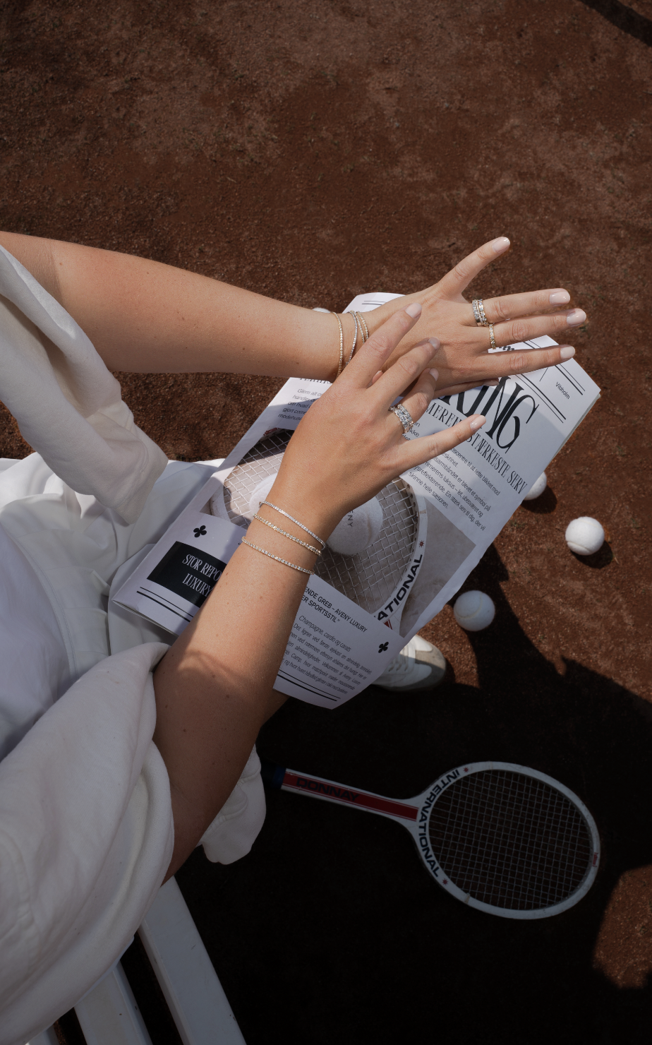 Nærbillede af en persons hænder med smykker, der læser en avis på en tennisbane med nogle golfbolde og en tennisracket til stede.
