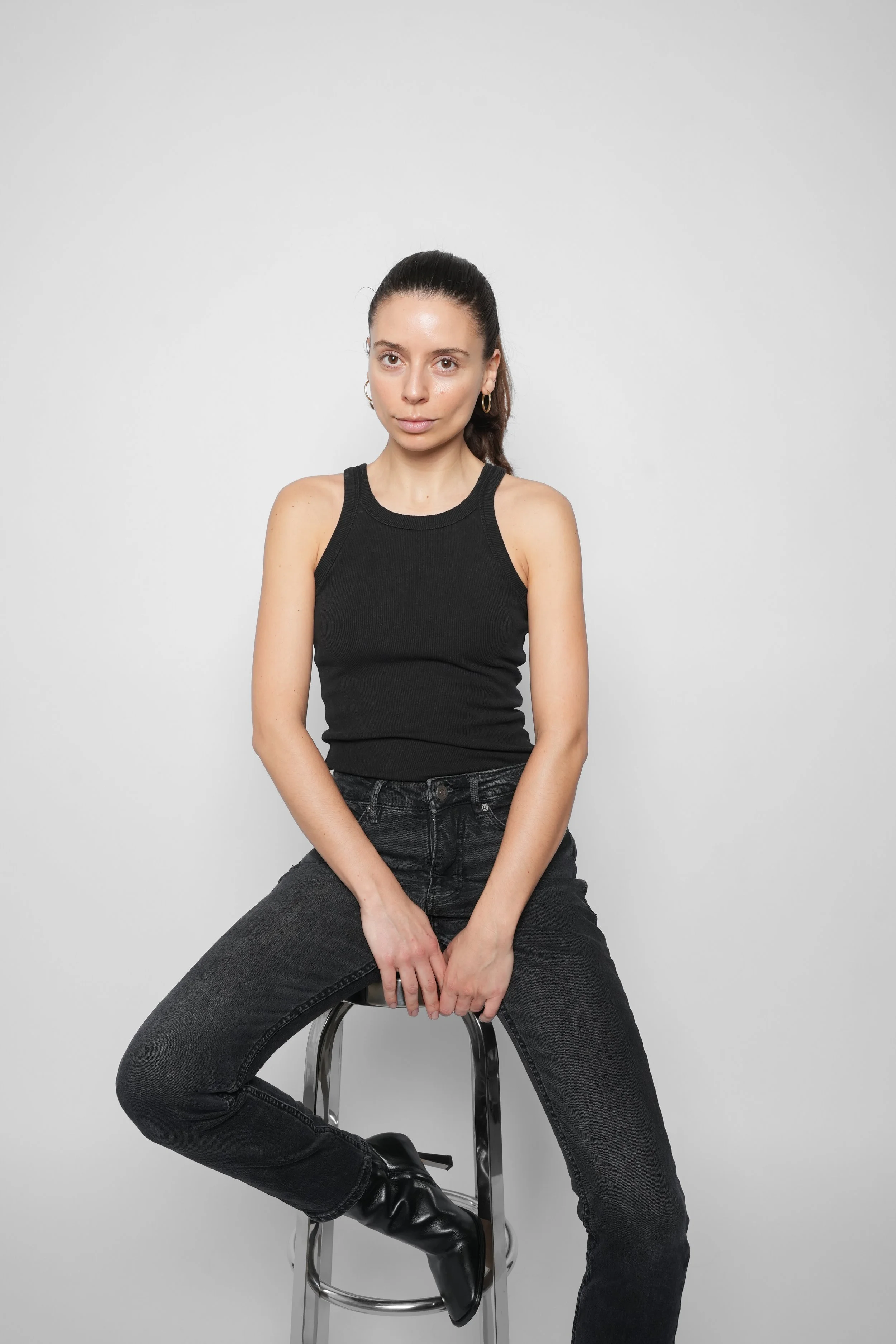 Kvinde i sort top og sorte jeans sidder på en barstol mod en hvid baggrund.