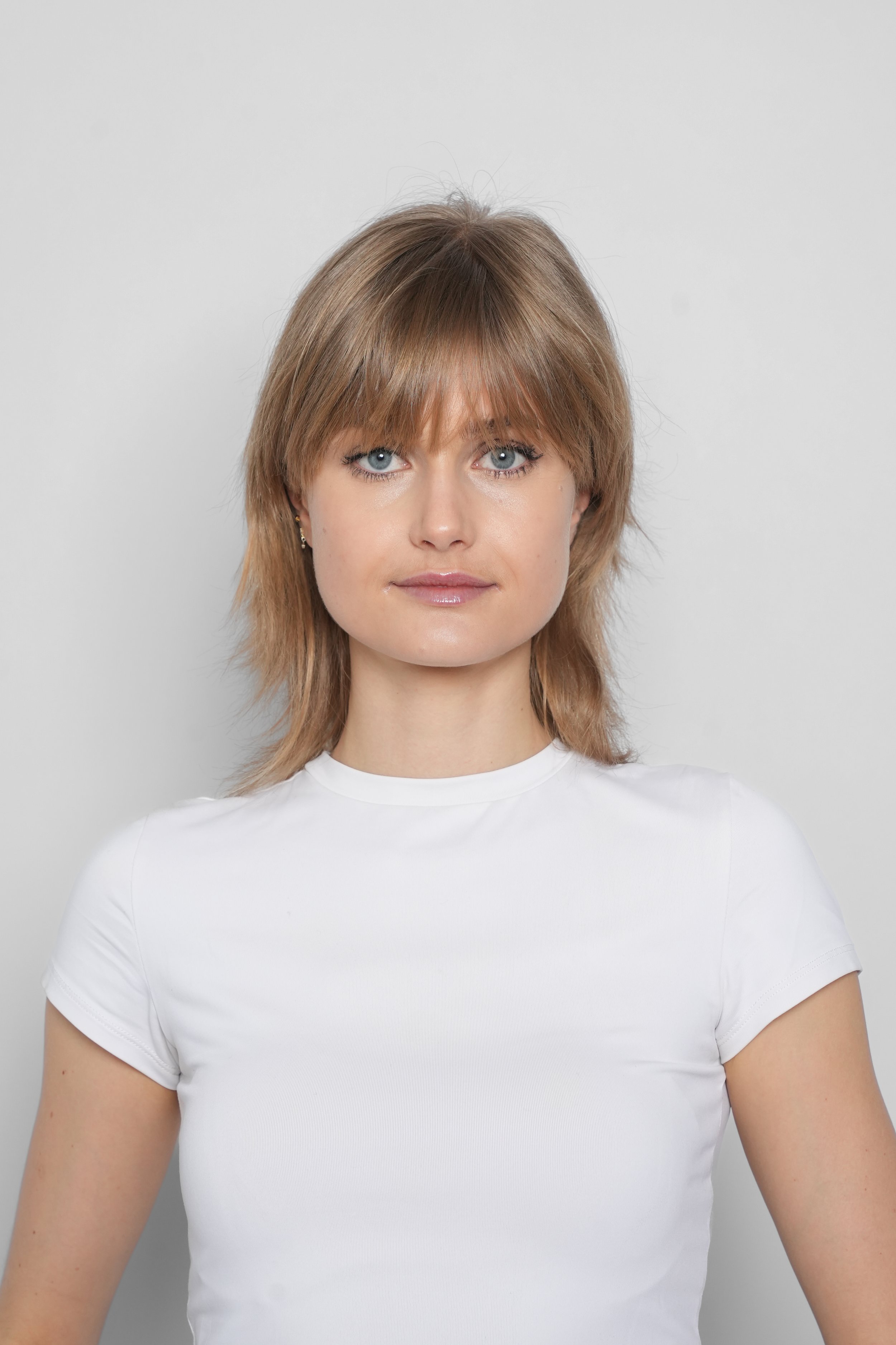 Portræt af en kvinde med brunt hår, lyse øjne, iført hvid t-shirt, foran en lys grå baggrund.