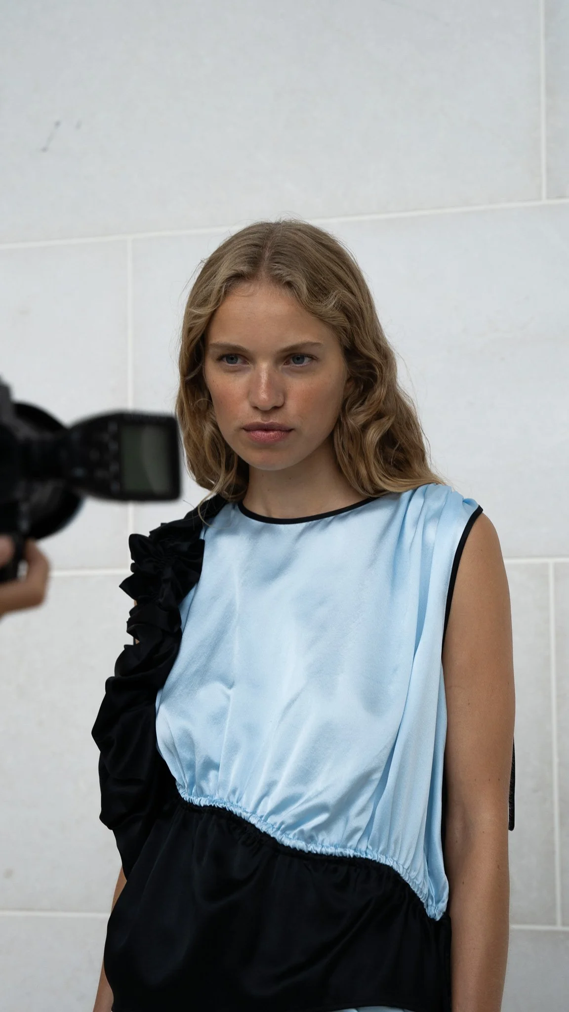 Kvinde med blond, krøllet hår poserer for kamera stående mod en hvid væg.