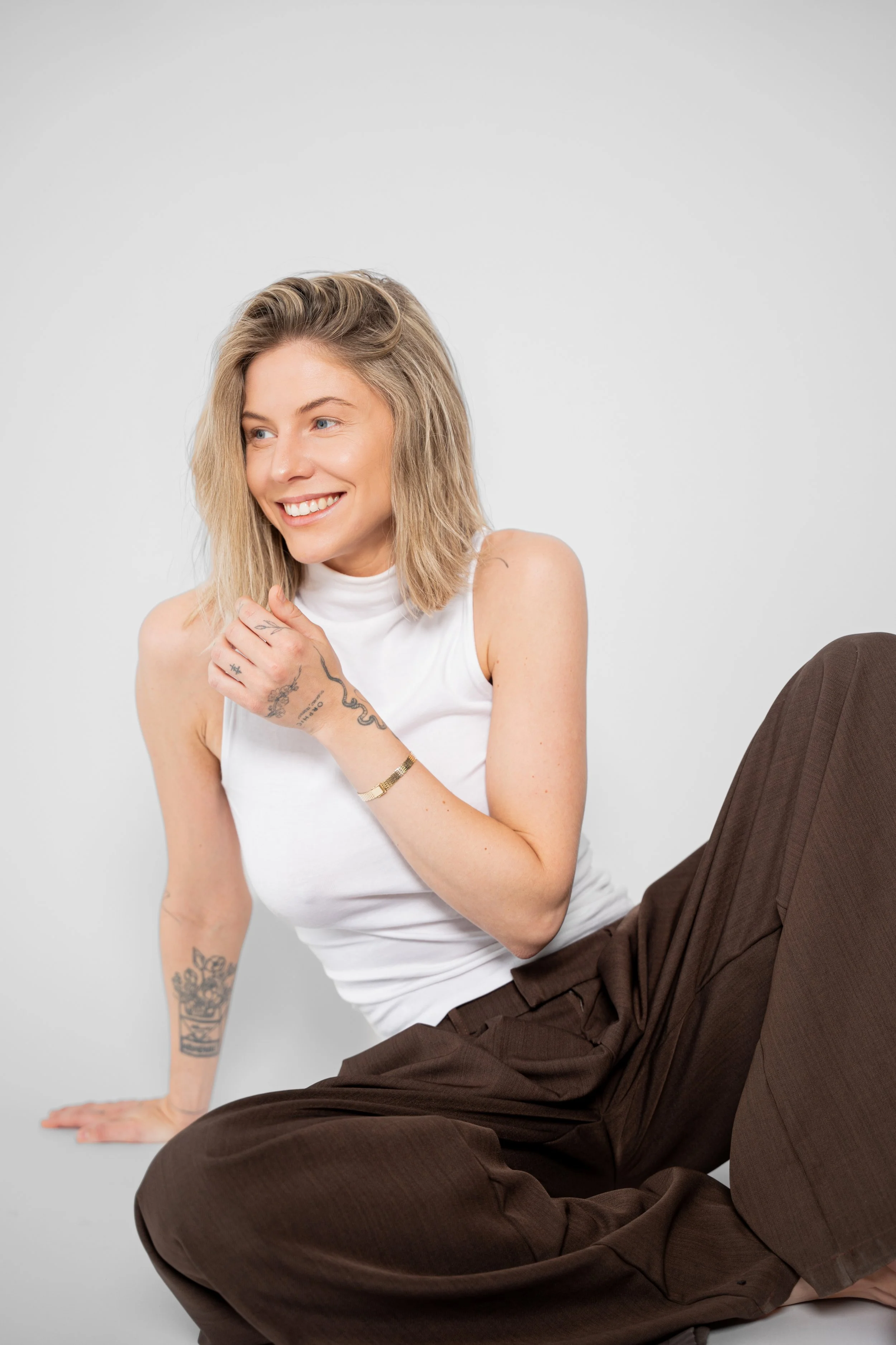 En smilende kvinde med blond hår, iført hvid top og brune bukser sidder på gulvet mod en hvid baggrund.