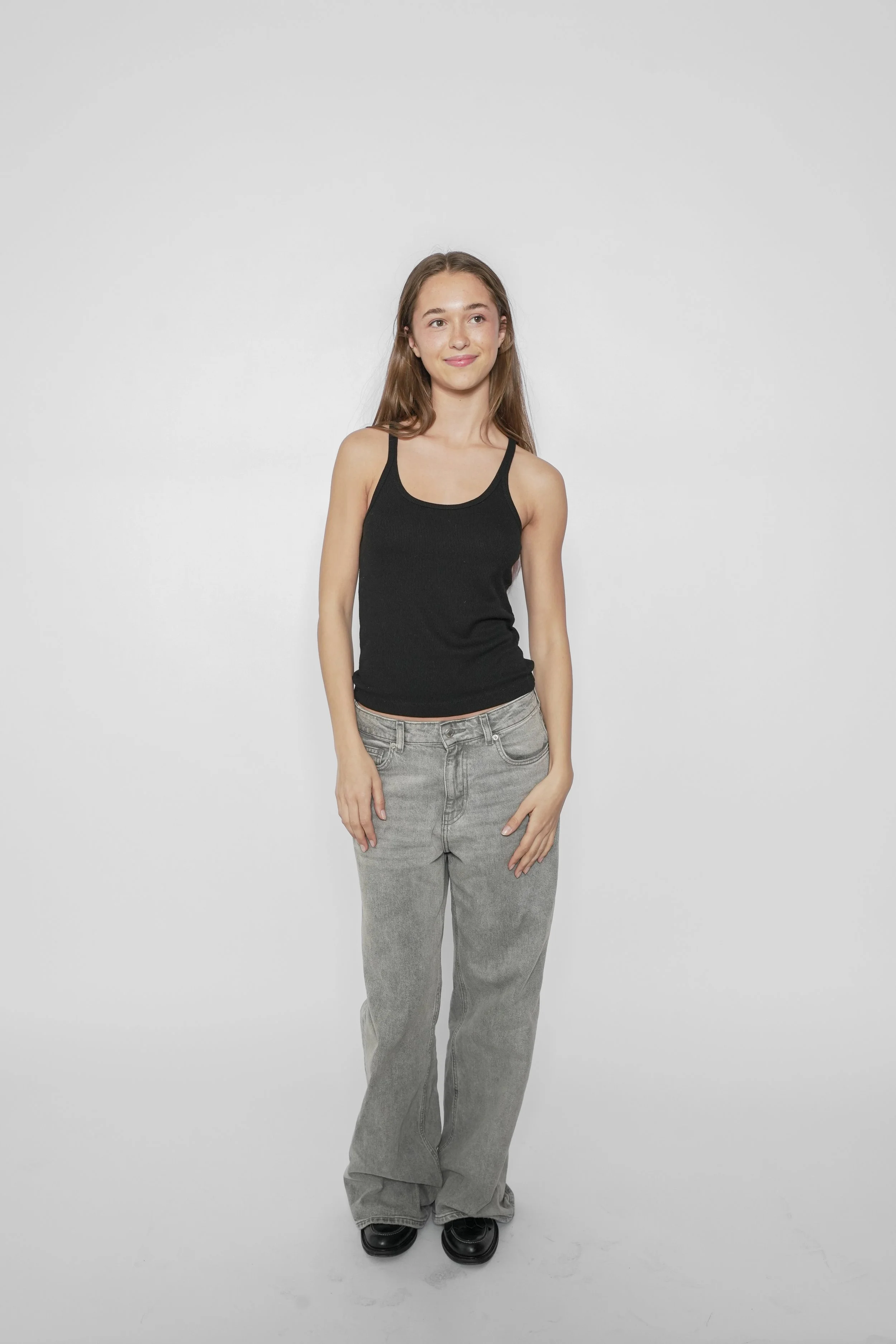 Ung pige iført sort tanktop og grå jeans står mod en hvid baggrund.