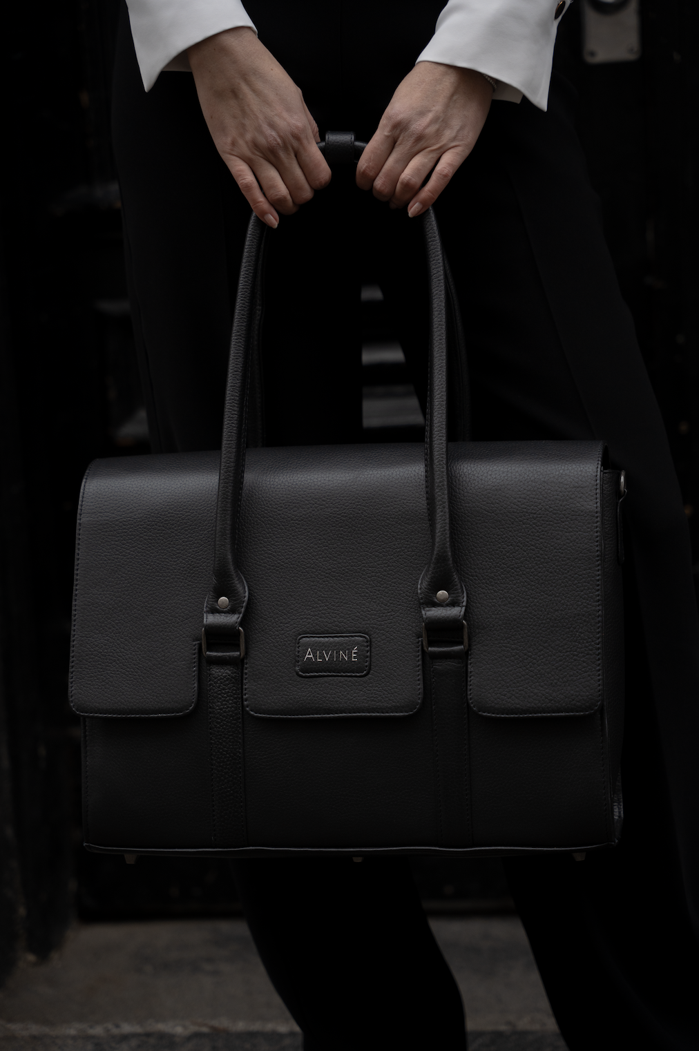 Person holder en sort taske med mærket 'Alviné', kun hænder og en del af kroppen er synlig.