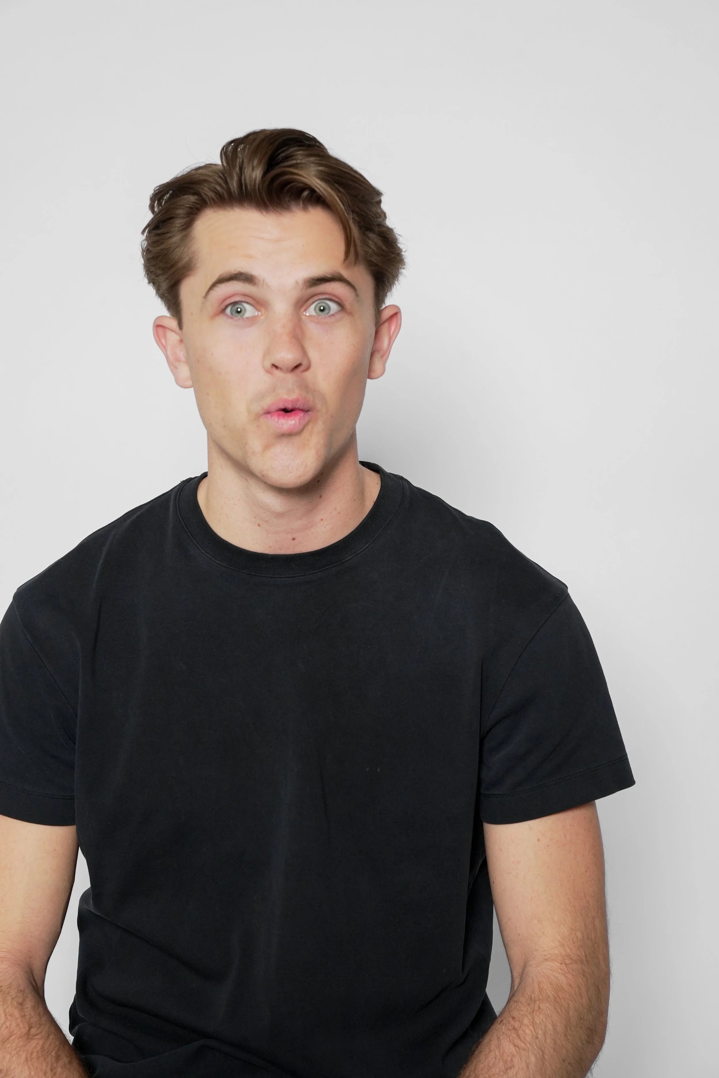 Ung mand med lys hud, brunt hår og blå øjne, der ser overrasket ud, iført sort T-shirt, foran en hvid baggrund.