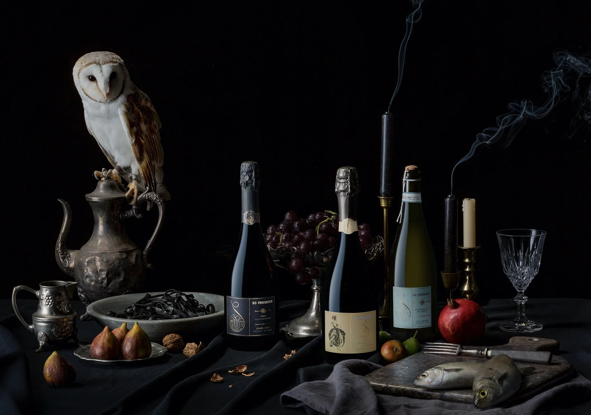 Still life of Prosecco, a pomegranate, figs, and fish on a dark table with a black background, candles, an owl figurine, and a glass.