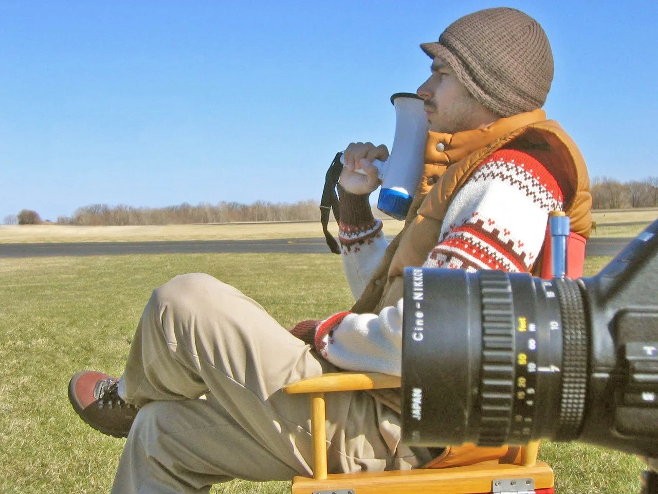 Frankie Latina sitting in a directors chair looking to the left holding a bullhorn with the lense of a movie camera in the foreground.