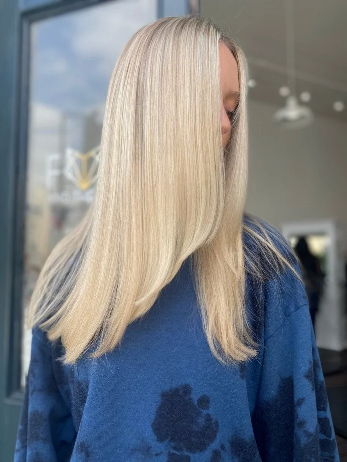 girl wearing blue stands outside hair salon modelling newly blonded hair that is long and layered