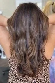woman standing in salon holds up hair showing freshly dyed light brown with caramel highlighted long wavy hair