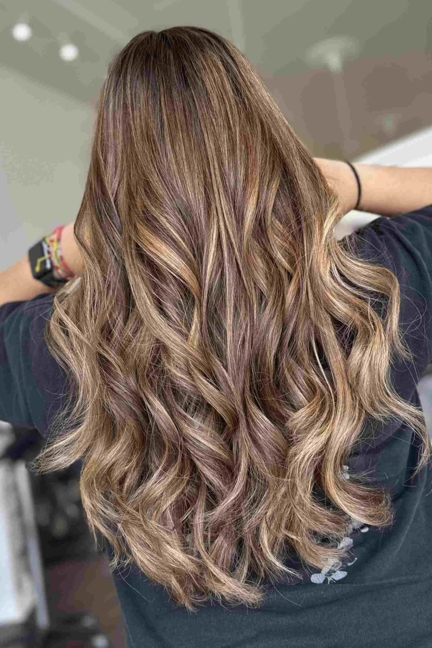 woman shows back of head displaying long curly brown hair with caramel highlights