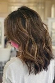 woman standing in salon shows back of  freshly dyed light brown hair with caramel highlights