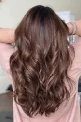 woman standing in salon holds up hair showing freshly dyed light brown with blonde highlighted long wavy hair