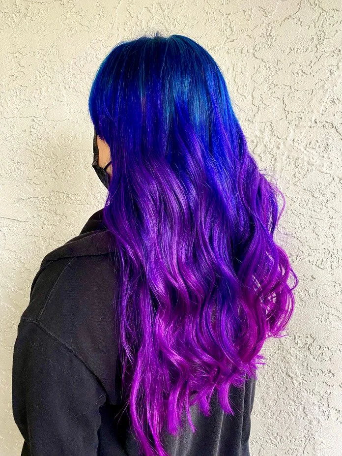 woman stands with back to camera showing off freshly colored vivid hair that is long and bright blue and purple