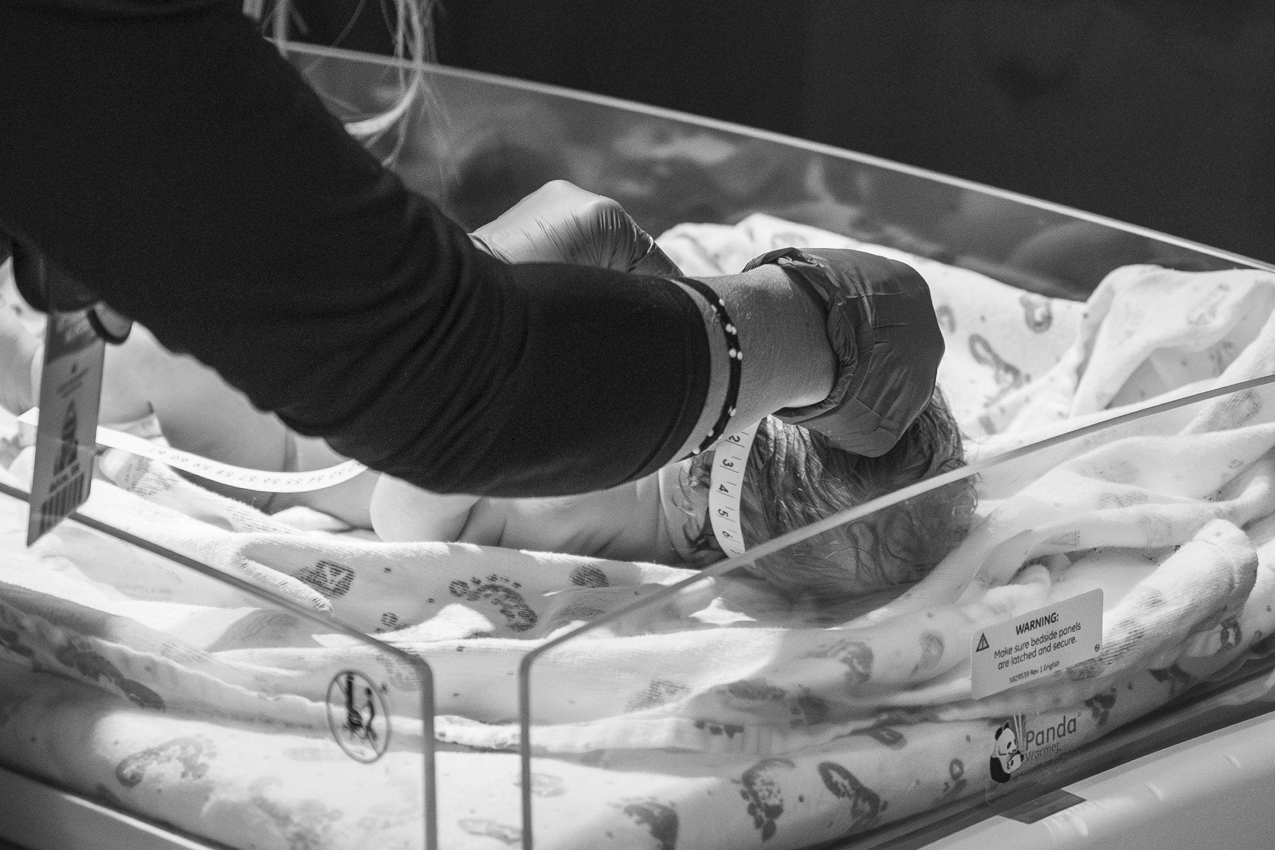 A medical professional in black clothing and gloves tending to a newborn baby in a clear bassinet, with a measuring tape around the baby's head.