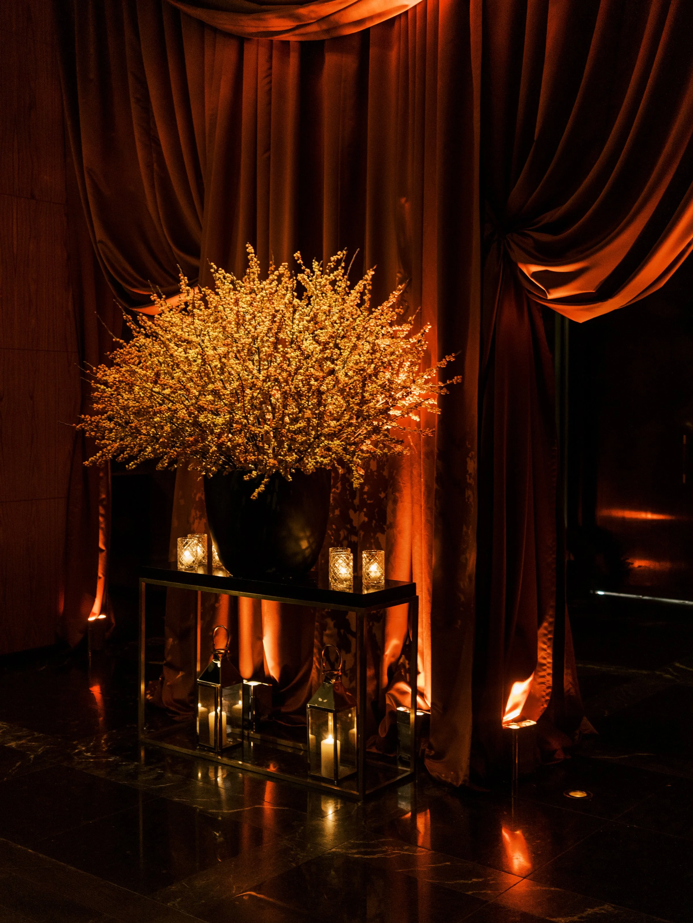 A dark room with large brown velvet curtains hanging behind a tall black vase with a bouquet of small white flowers. Small lanterns with candles inside are on a black table below the vase. The ambient lighting creates a warm glow.
