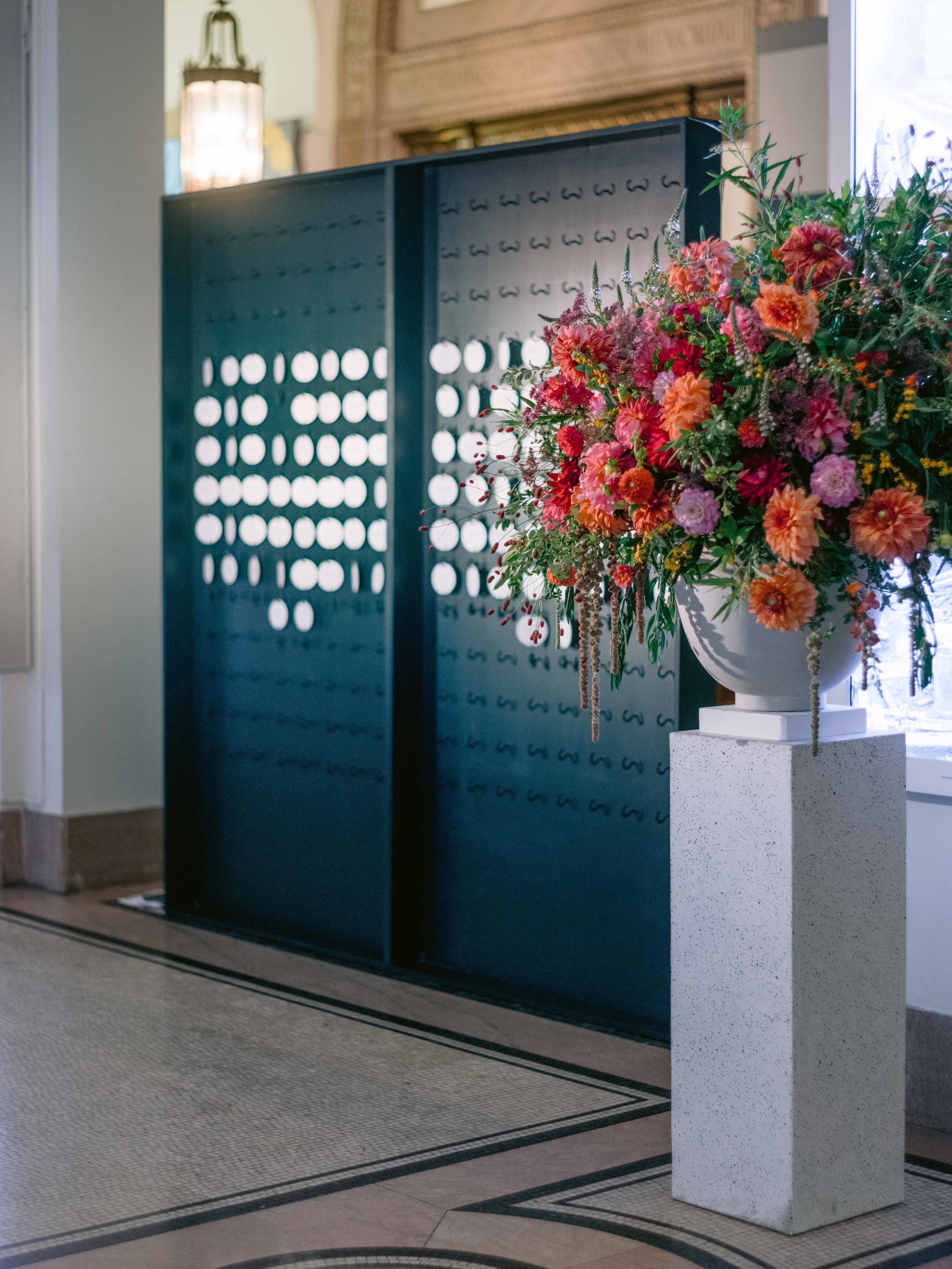 A large bouquet of pink, orange, and purple flowers in a white vase on a white speckled pedestal inside a building.