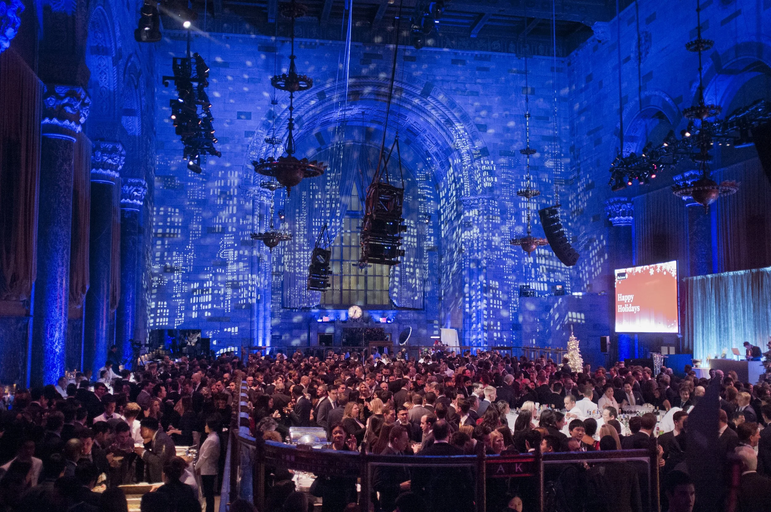 A large indoor event with many people seated at tables, with blue lighting and a decorative, ornate architectural interior, including stained glass windows and chandeliers.