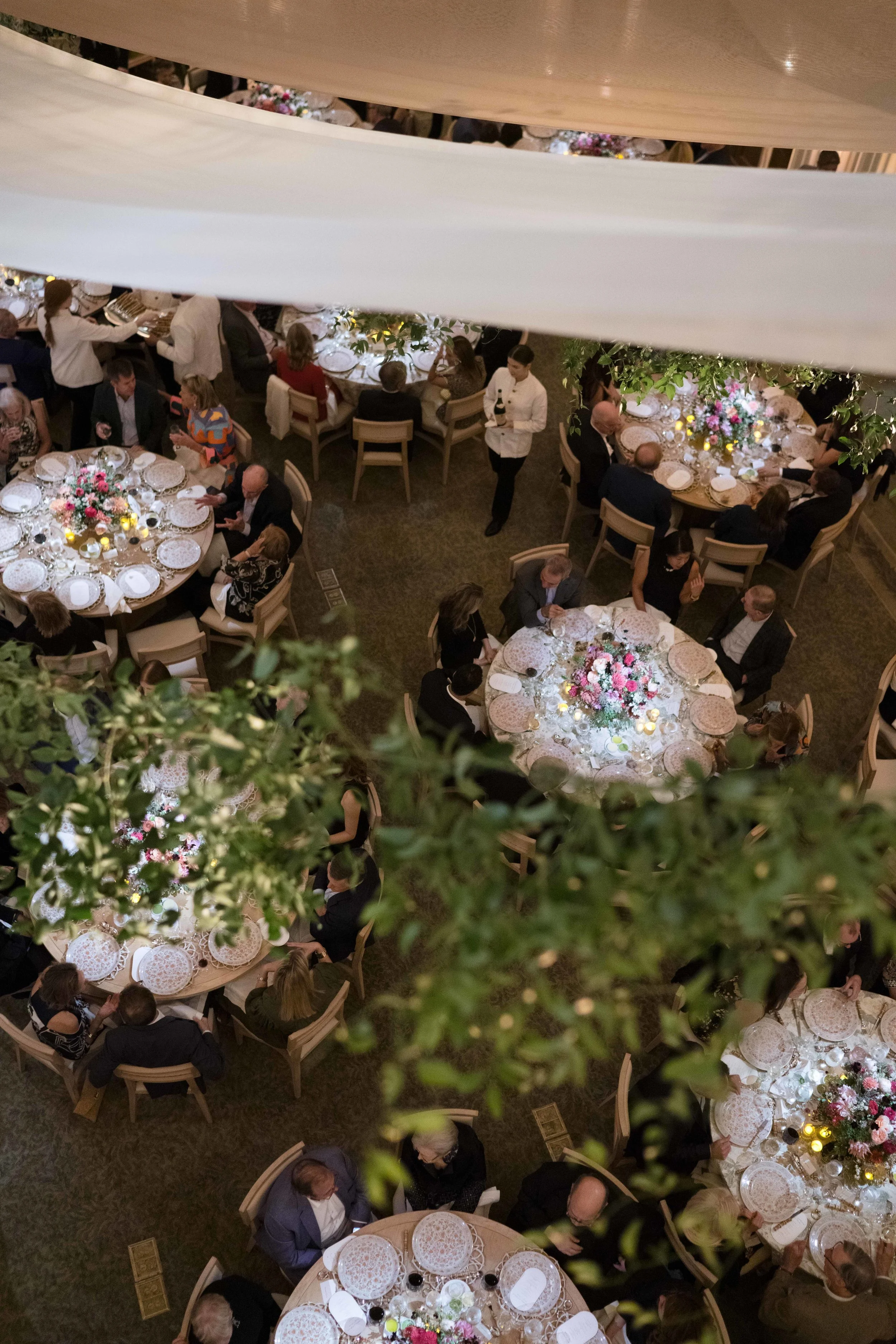A banquet hall filled with round tables set for a formal event, decorated with floral centerpieces and lit with candles, viewed from above through decorative drapes and greenery.