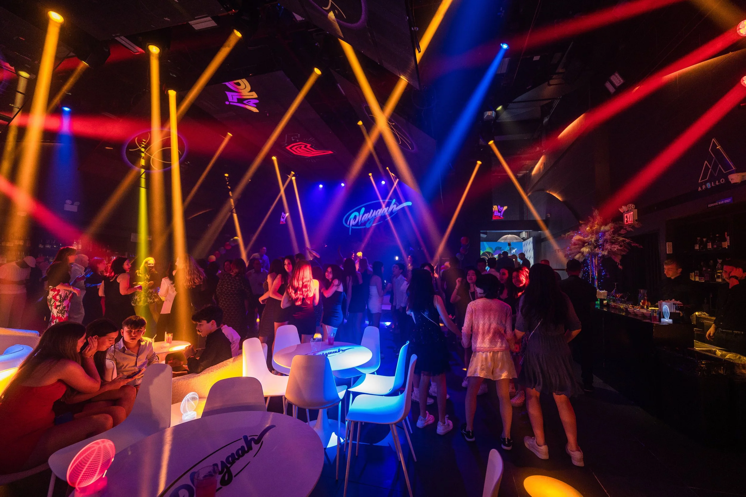 A lively nightclub with colorful laser lights and a crowd of young people dancing and socializing. Neon signs and decorations add to the energetic atmosphere.