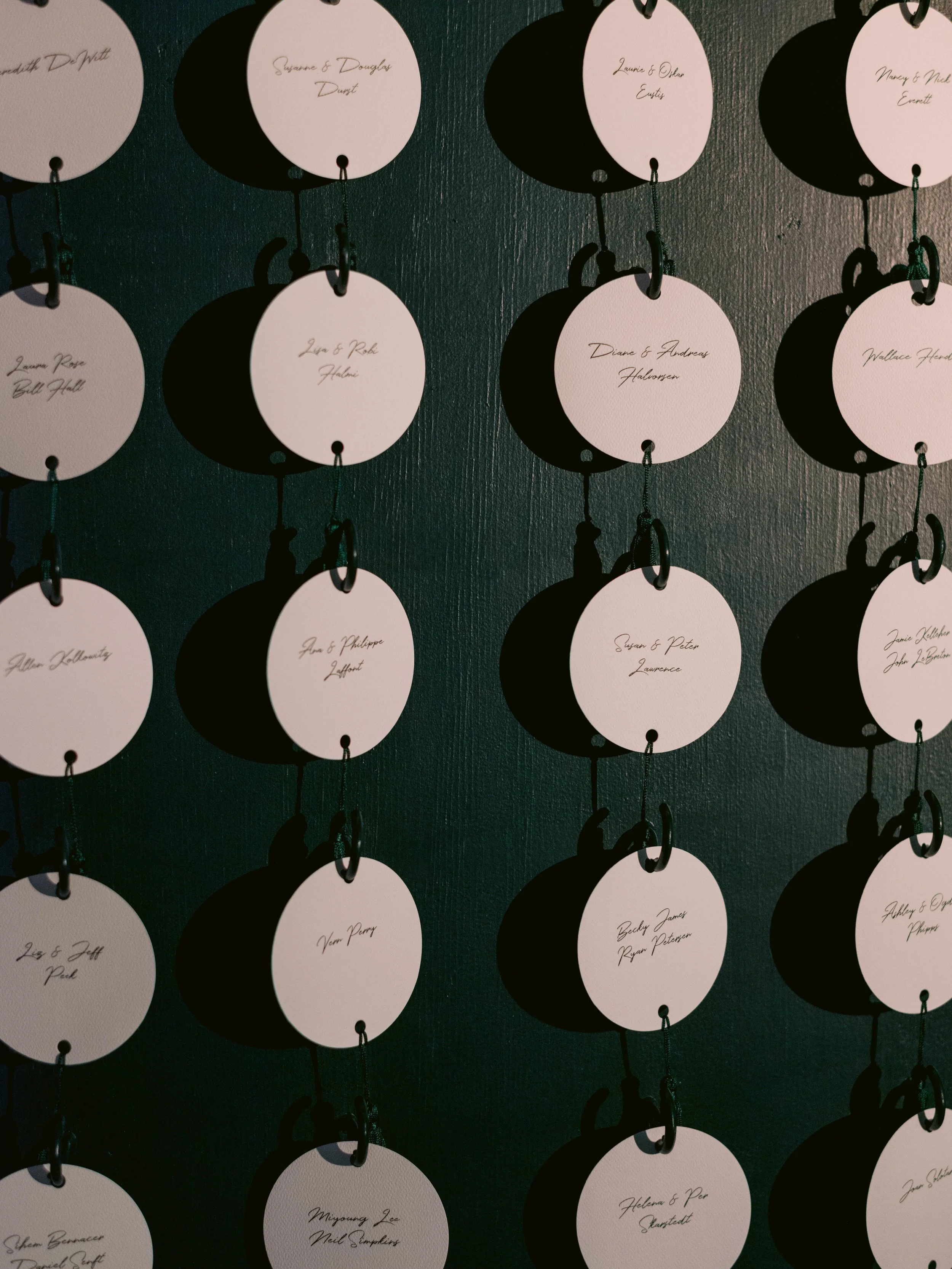 Multiple white circular tags with handwritten names and pairs hanging on black hooks against a black textured wall.