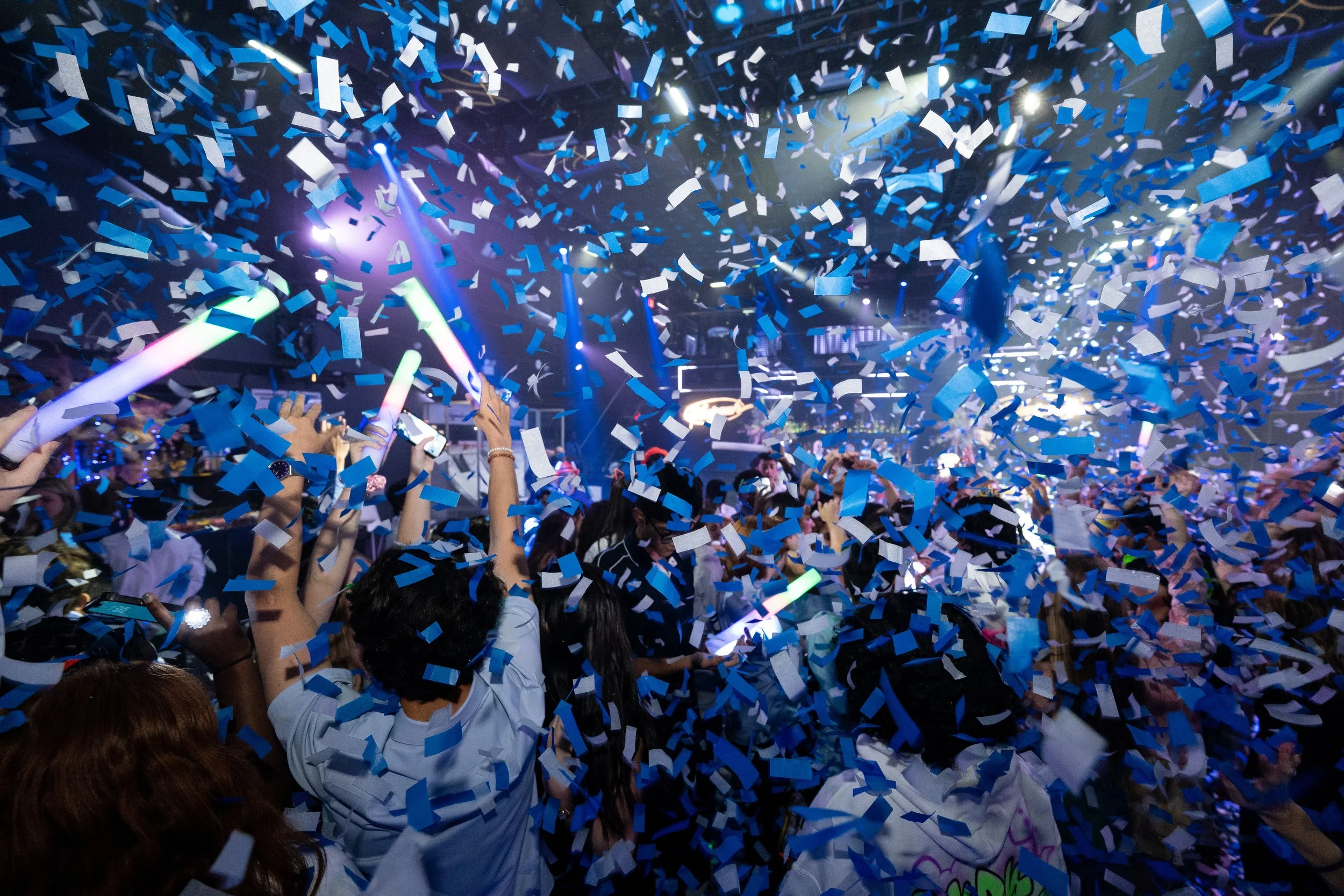 A lively crowd at a concert or event, with blue and white confetti falling from the ceiling, and people holding glow sticks and taking pictures.