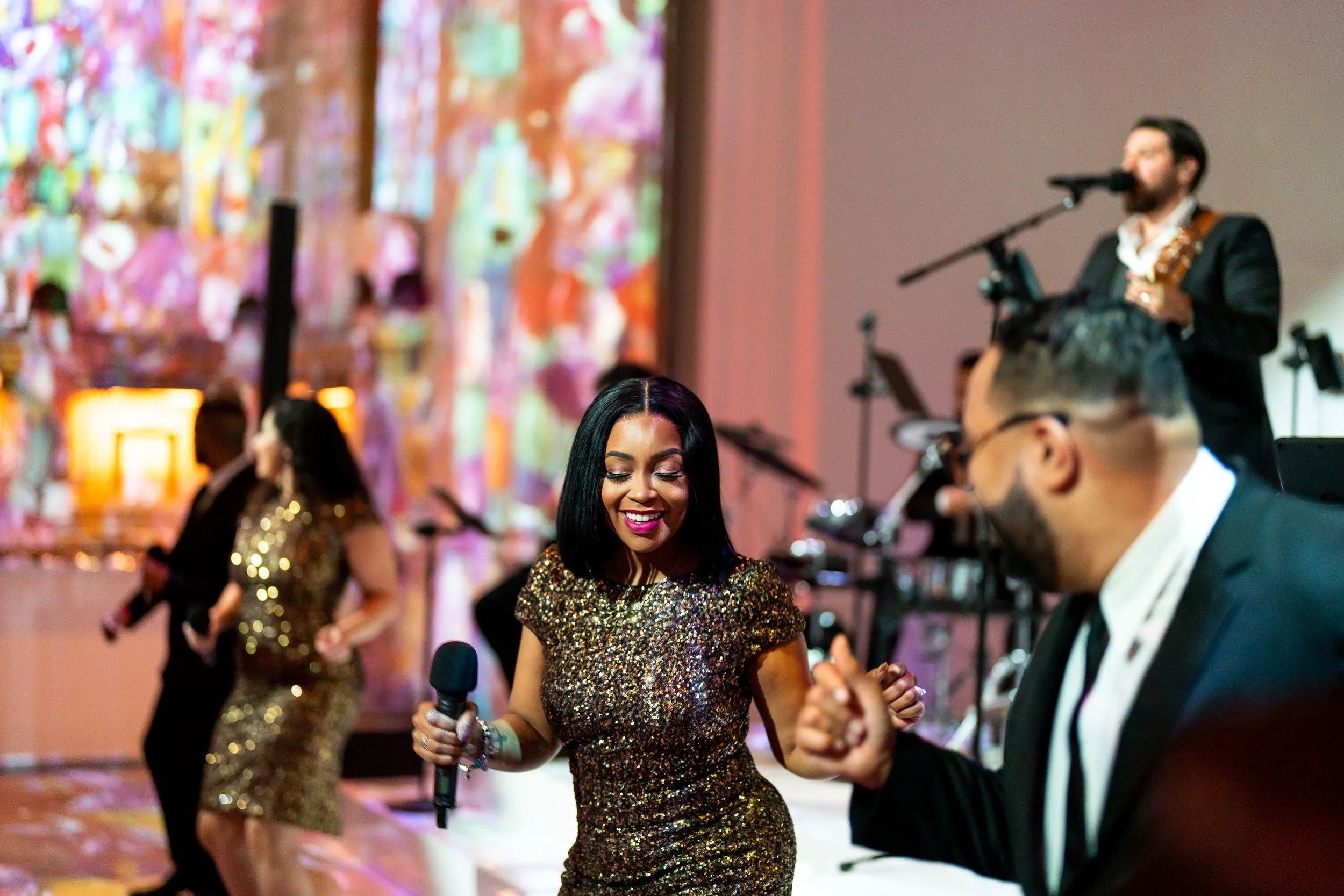 A woman in a sparkling gold dress holds a microphone, dancing with a man in a tuxedo at a lively party or celebration. In the background, other guests are dancing, and colorful abstract artwork decorates the wall.
