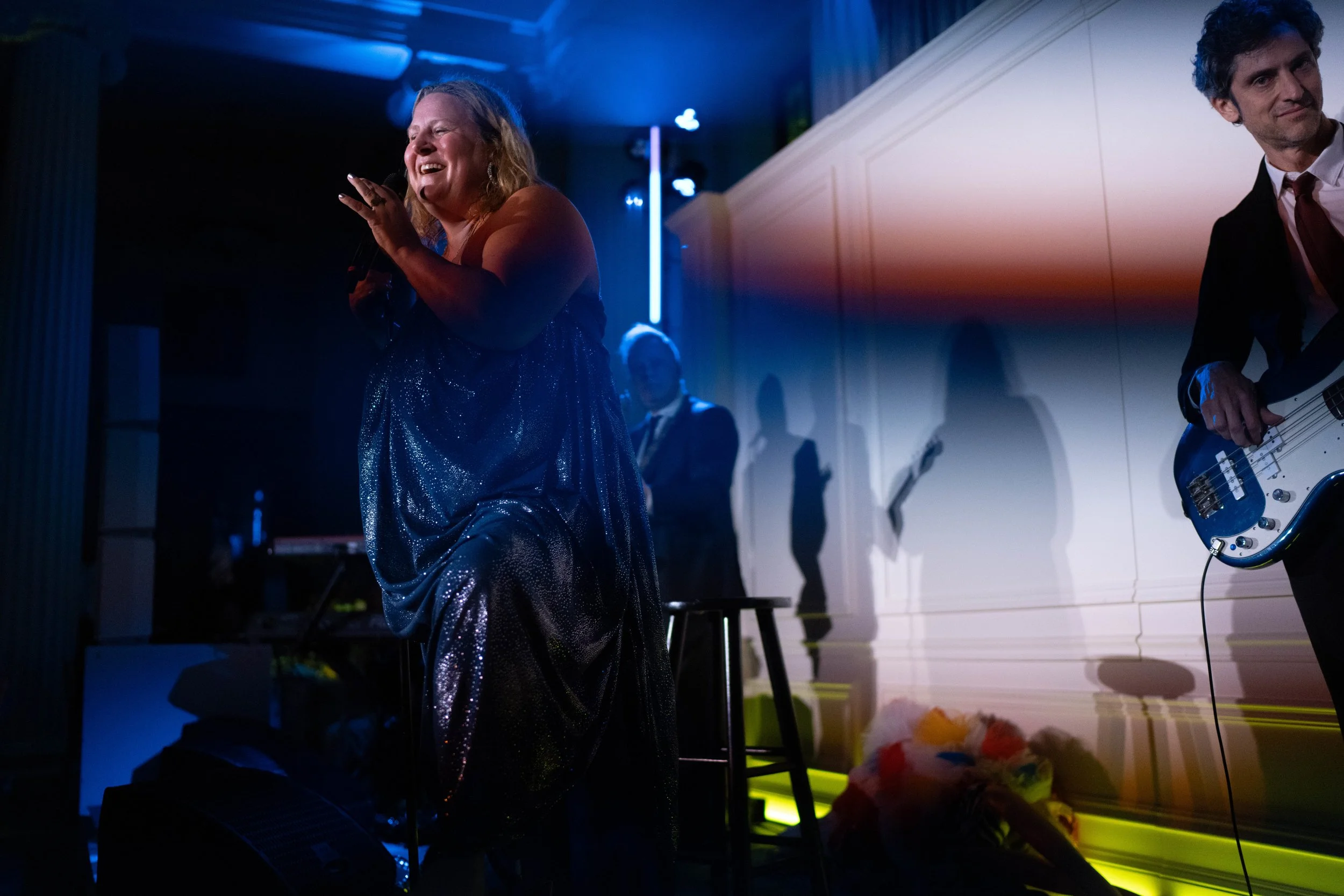 Woman singing into a microphone on stage, with band members playing instruments and colorful lighting in the background.