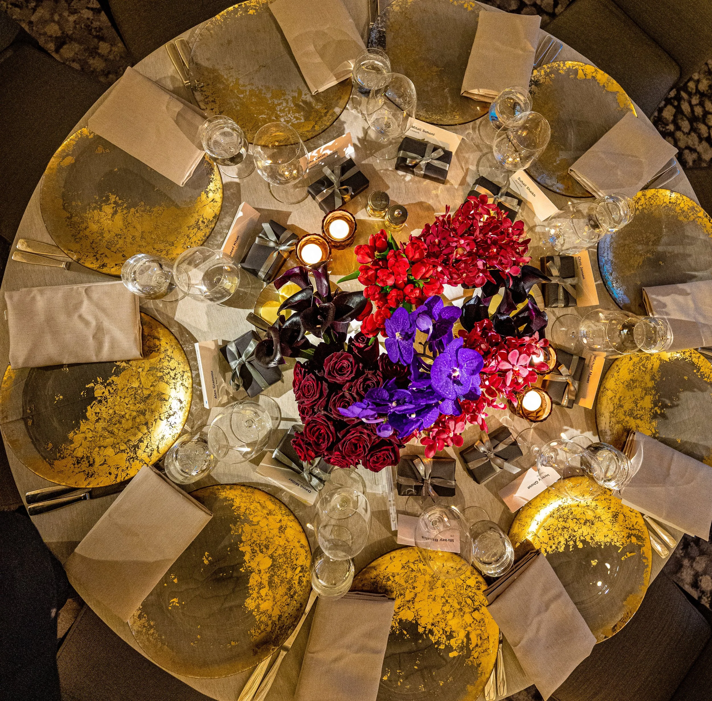 A top-down view of a round dining table set for a celebration with gold-accented plates, wine glasses, wrapped gifts, candles, and a bouquet of vibrant purple, pink, and red flowers.